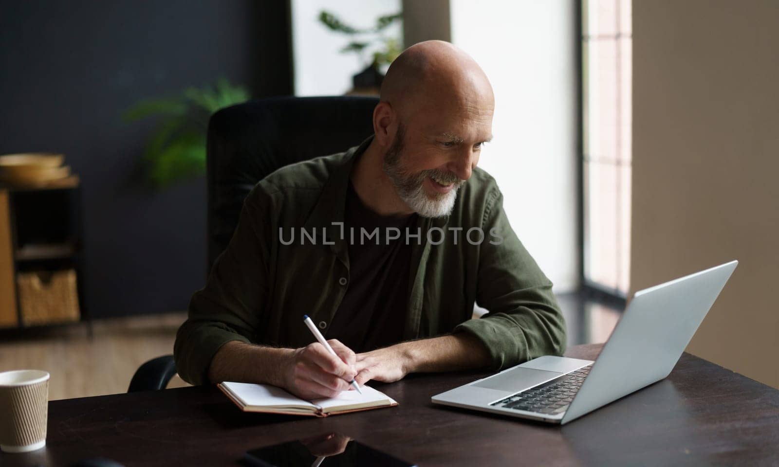 Smiling mid-aged man focused and productive state. He seated at desk indoors, engrossed work. With notepad in front, he diligently writing down notes, while simultaneously looking at computer screen. . High quality photo
