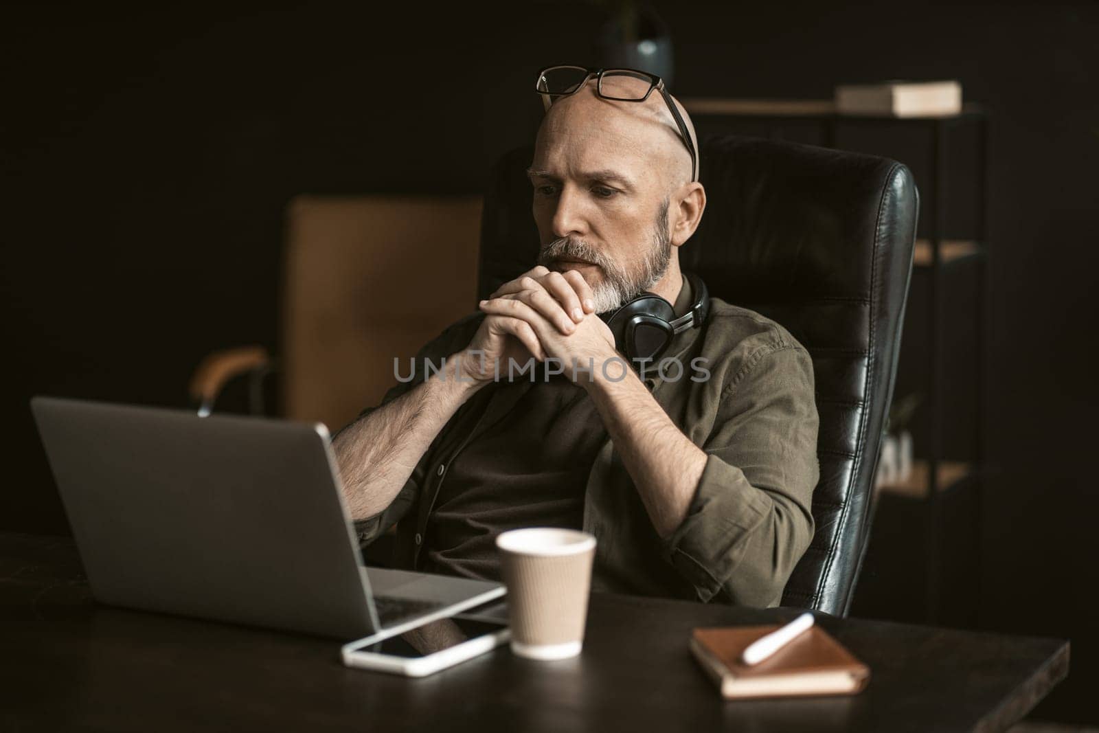 Dedicated elderly worker in loft office environment. With puzzled expression on his face, he sits attentively in front of a computer, deeply engaged in solving a complex problem. . High quality photo