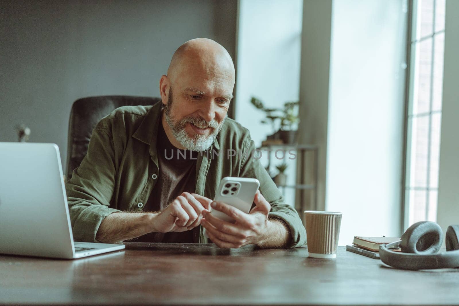 Mature old man comfortably seated home, actively engaged in texting message on phone. Desk adorned with laptop, headphones, and cup of coffee, indicating setup conducive to work and relaxation. by LipikStockMedia