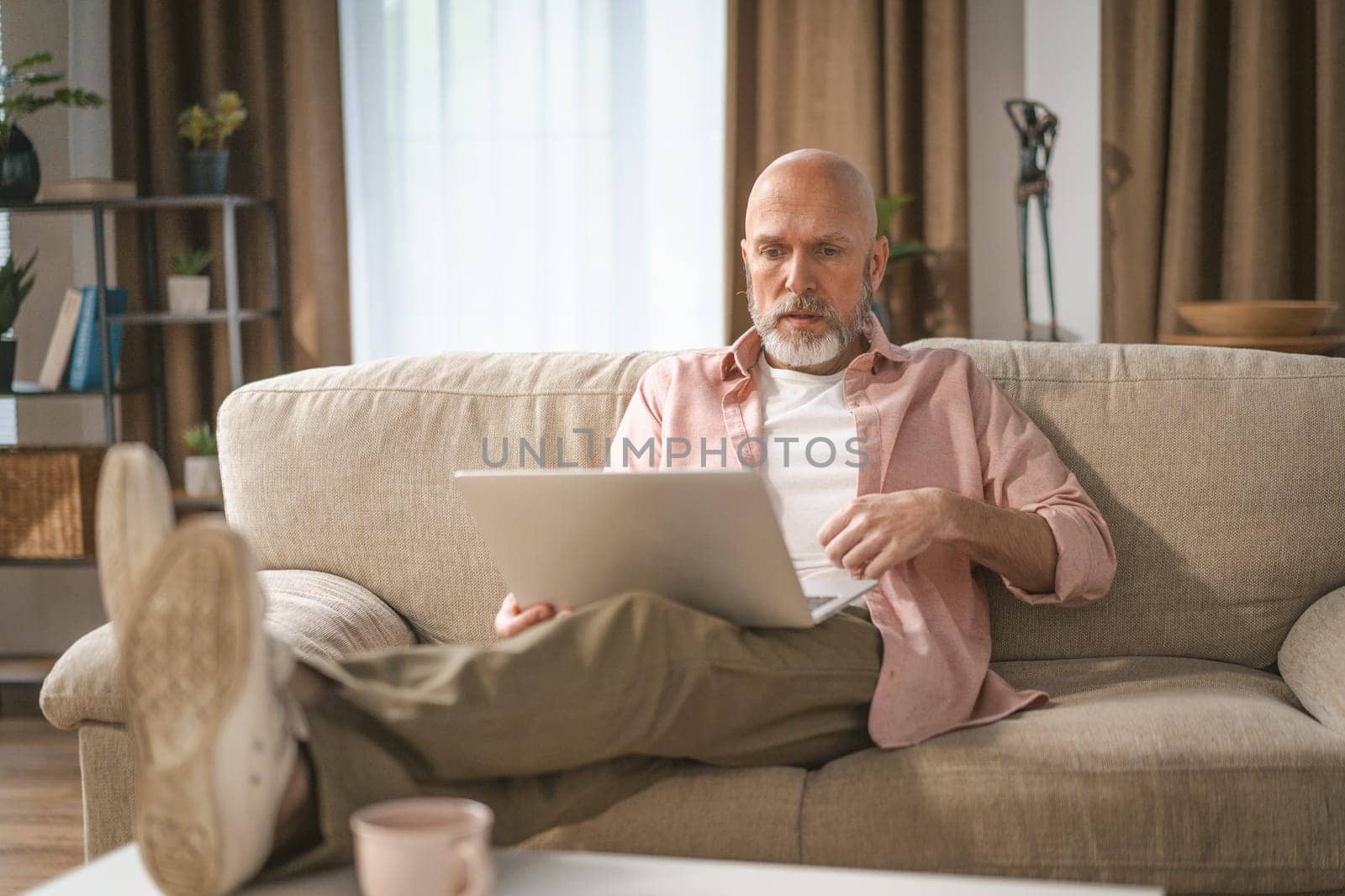 Senior man with distinguished silver beard depicted enjoying time home. He seated comfortably on sofa, engrossed in watching internet on his laptop. High quality photo