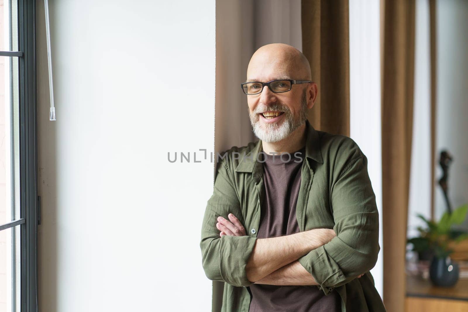 Happy old age as a mature man radiates positivity from the comfort of his own home. Positioned near a window, he smiles warmly towards the camera, emanating a sense of contentment and joy. . High quality photo