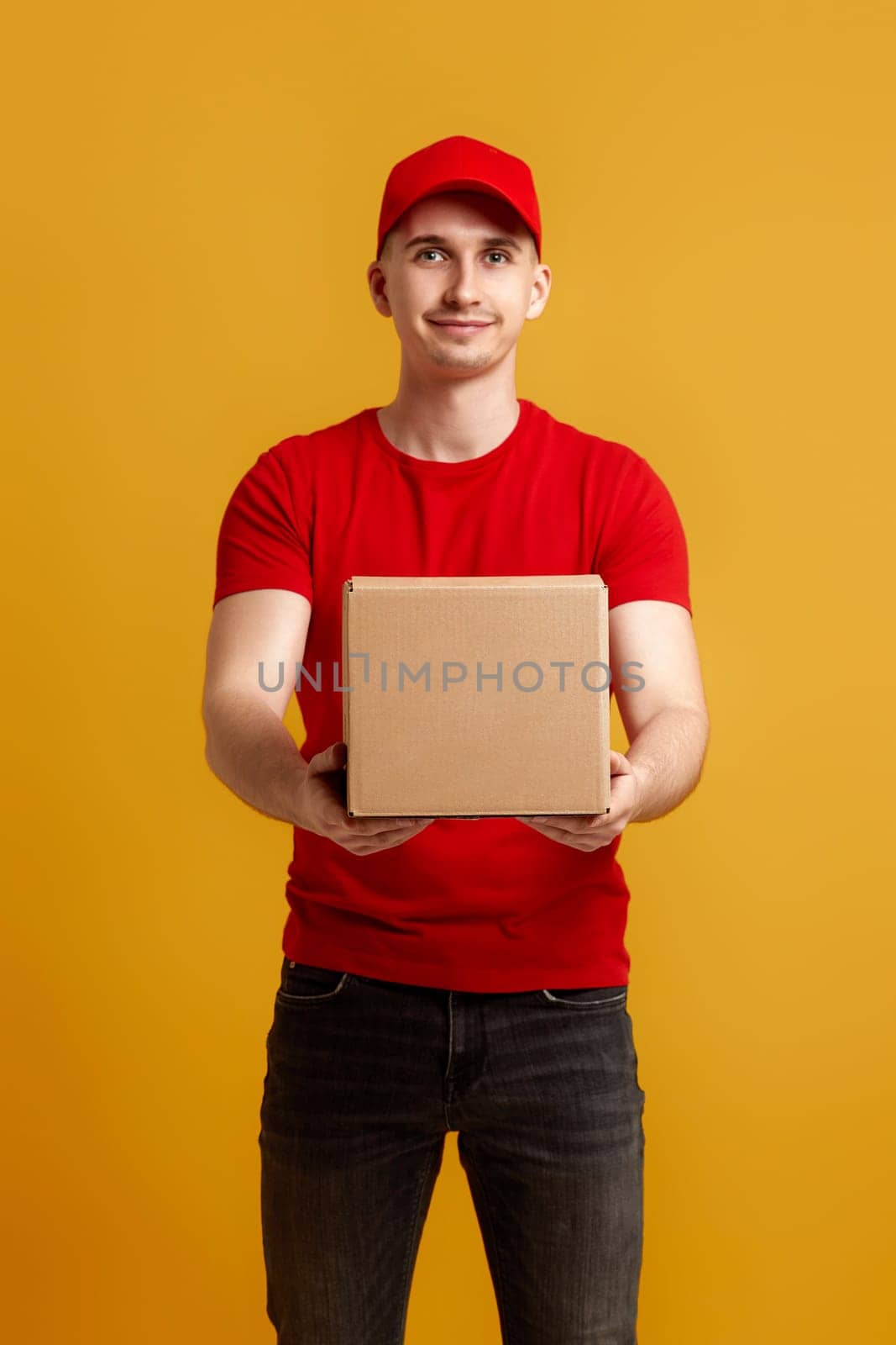 Delivery man employee in red cap, red t-shirt holding cardboard box isolated on yellow background