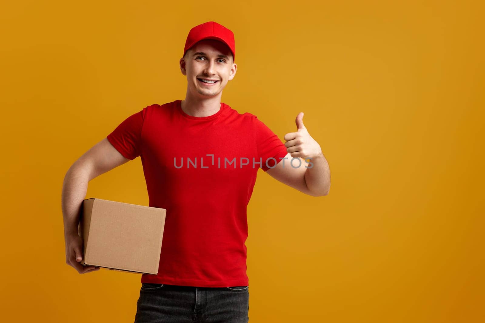 smiling delivery man employee in red cap, t-shirt holding cardboard box isolated on orange background