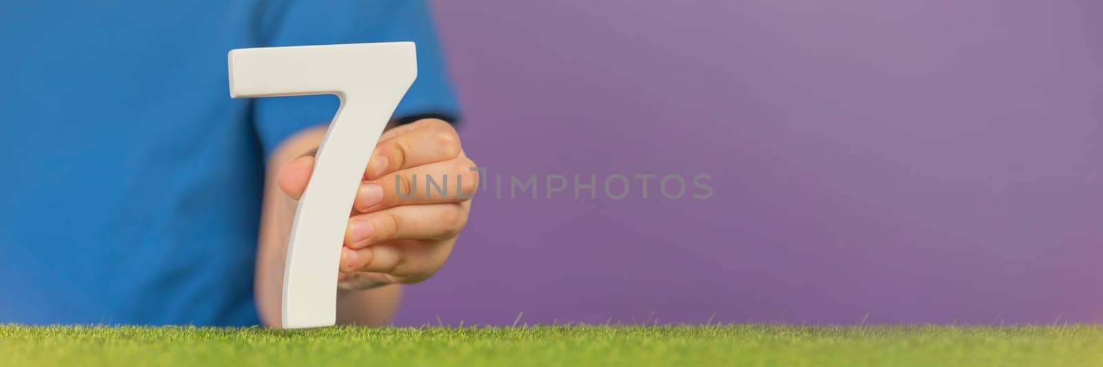 Number 7 in hand. A hand holds a white number seven on green grass and a purple background, seven percent per annum. World Health Day. April 7th. Holiday concept. by SERSOL