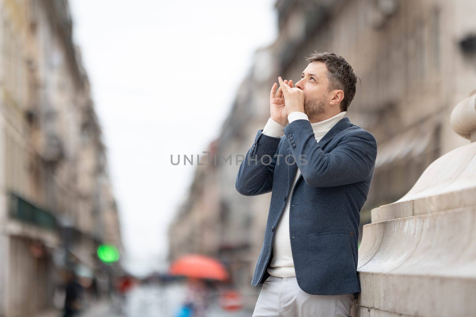 A man in a blue jacket plays the harmonica on the street looking up. Mid shot