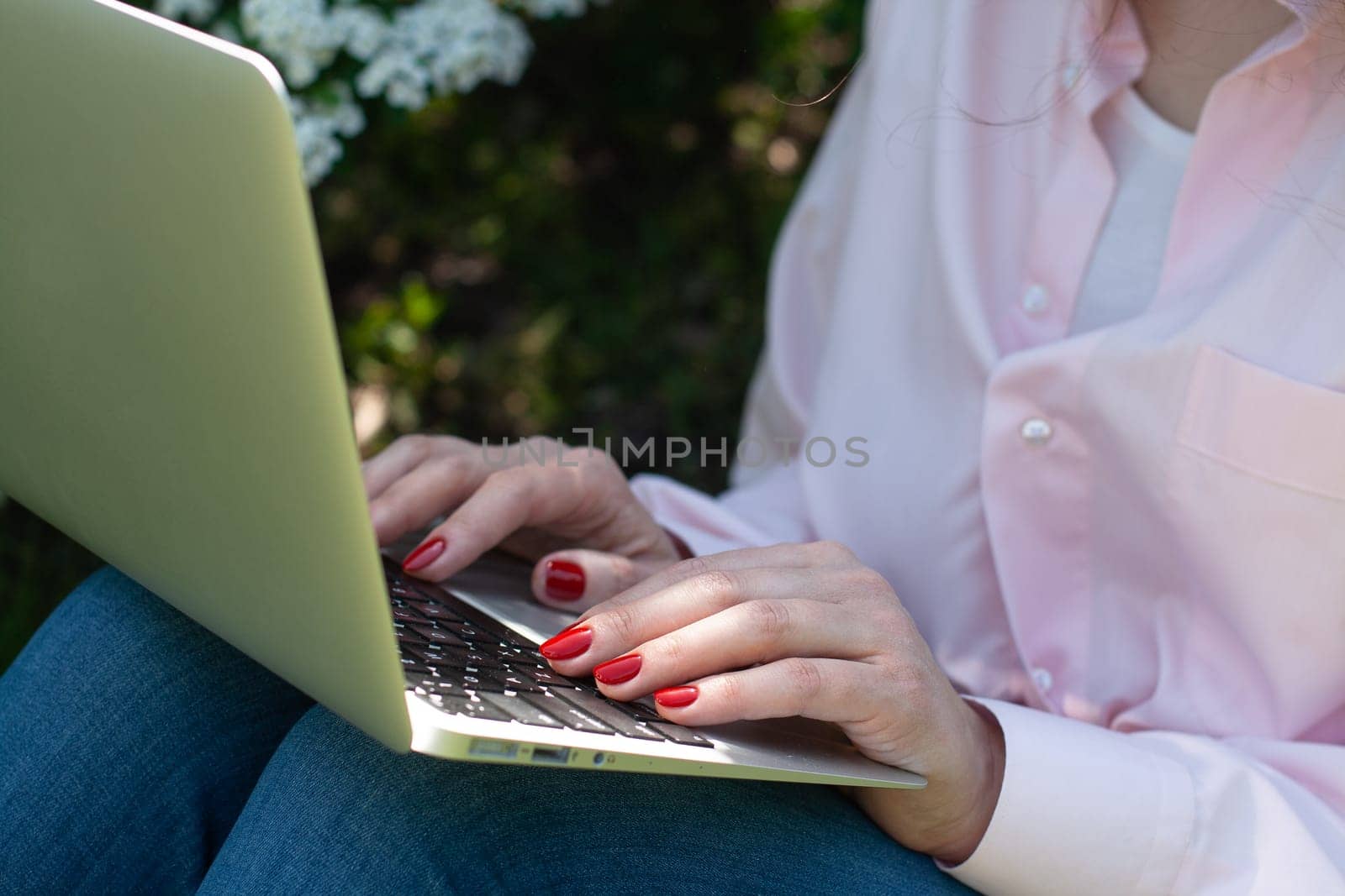 A girl in a pink shirt sits in the garden on the grass with a laptop, hands with a red manicure