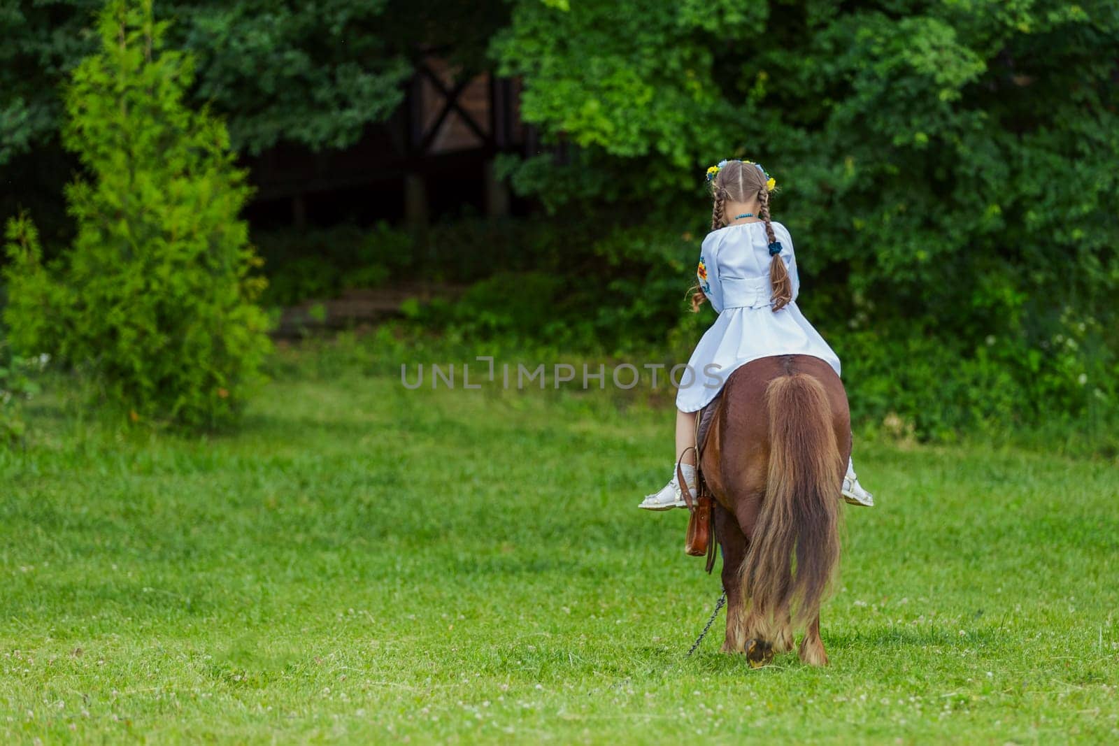 A little girl in the Ukrainian national costume rides a pony on the lawn