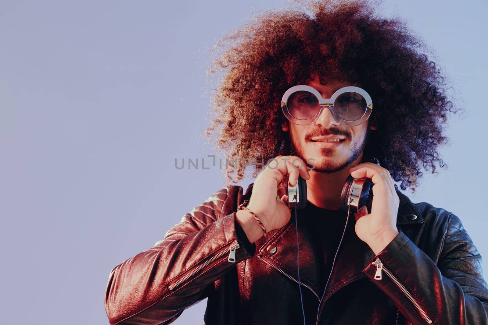Portrait of a stylish man with curly hair with glasses and headphones on a blue background multinational, colored light, black leather jacket trend, modern concept. High quality photo