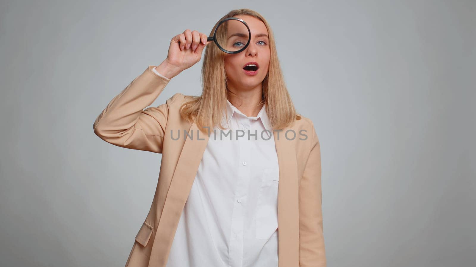 Investigator researcher scientist office woman holding magnifying glass near face, looking into camera with big zoomed funny eyes, searching analysing. Business girl isolated on gray studio background
