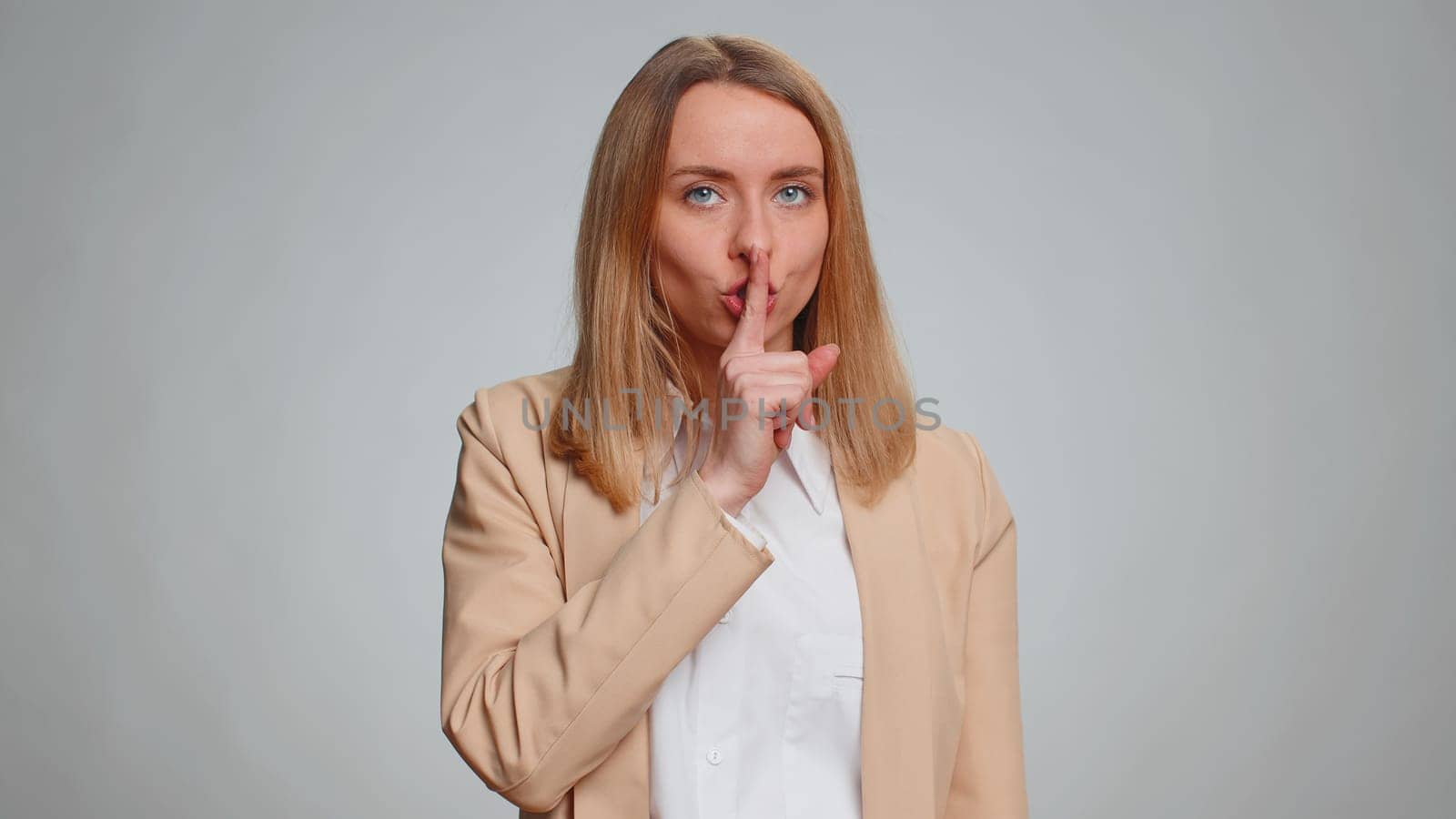 Shh be quiet please, keep my secret. Portrait of young businesswoman presses index finger to lips makes silence gesture sign do not tell anything. Female girl in suit. Pretty woman on gray background