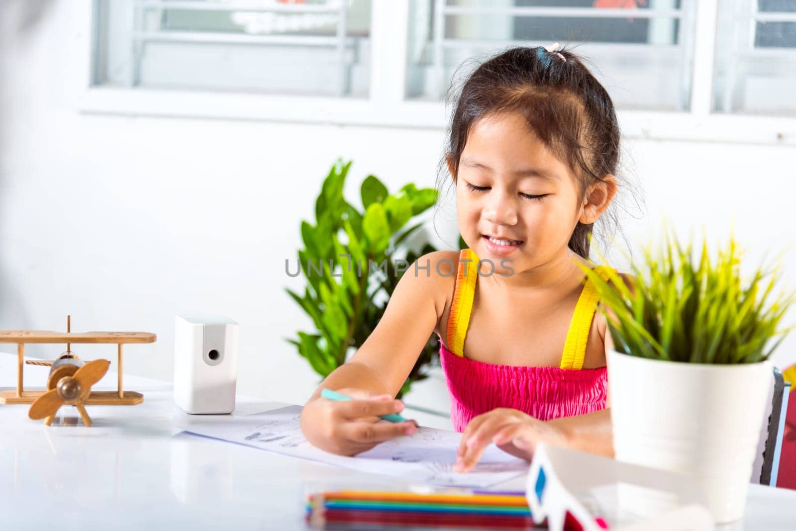 Child little girl drawing cartoon on paper before paint the color, Asian cute kid preschooler sitting on table smiling she draw and writing a picture with pencil at home to learning arts homework