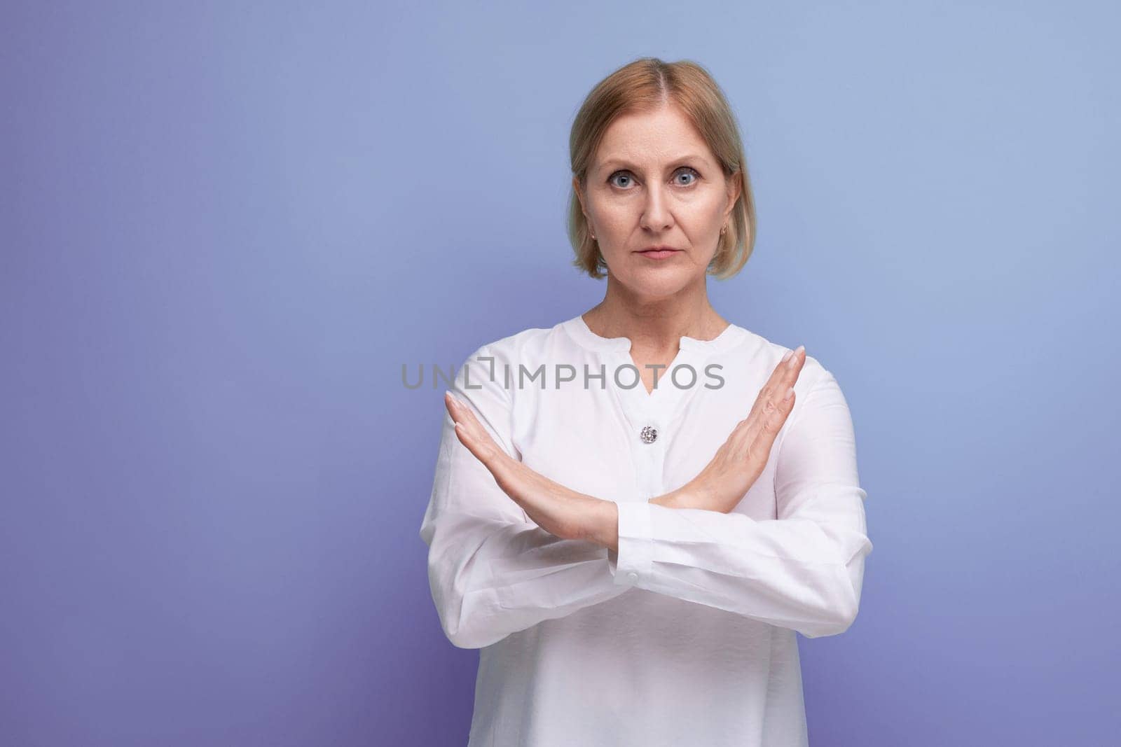 serious blond middle aged woman in white blouse with hand cross gesture on studio background.