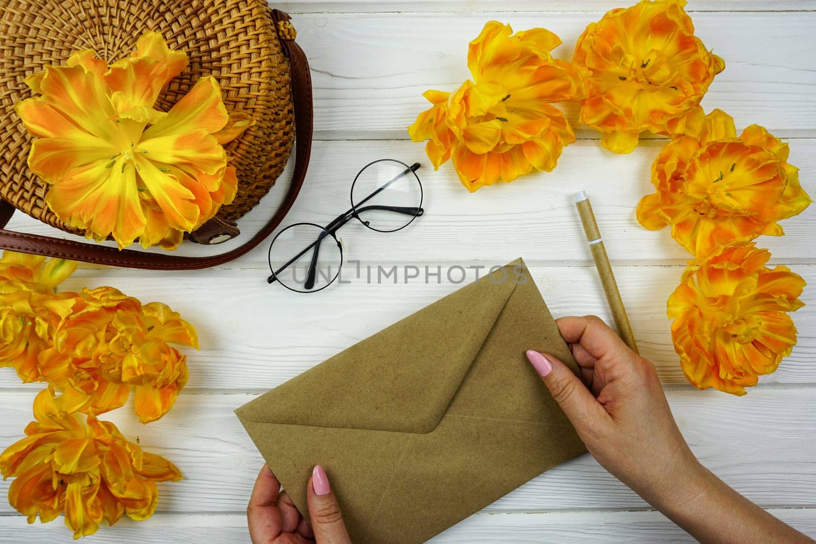 Women's hands are holding a craft envelope. On a white wooden table there is a wicker bag and yellow terry tulips. by Spirina