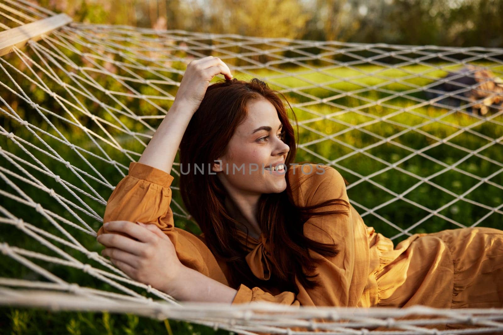a happy woman is resting in a mesh hammock with her head resting on her hand, smiling happily looking away, enjoying a warm day in the rays of the setting sun, lying in an orange dress by Vichizh