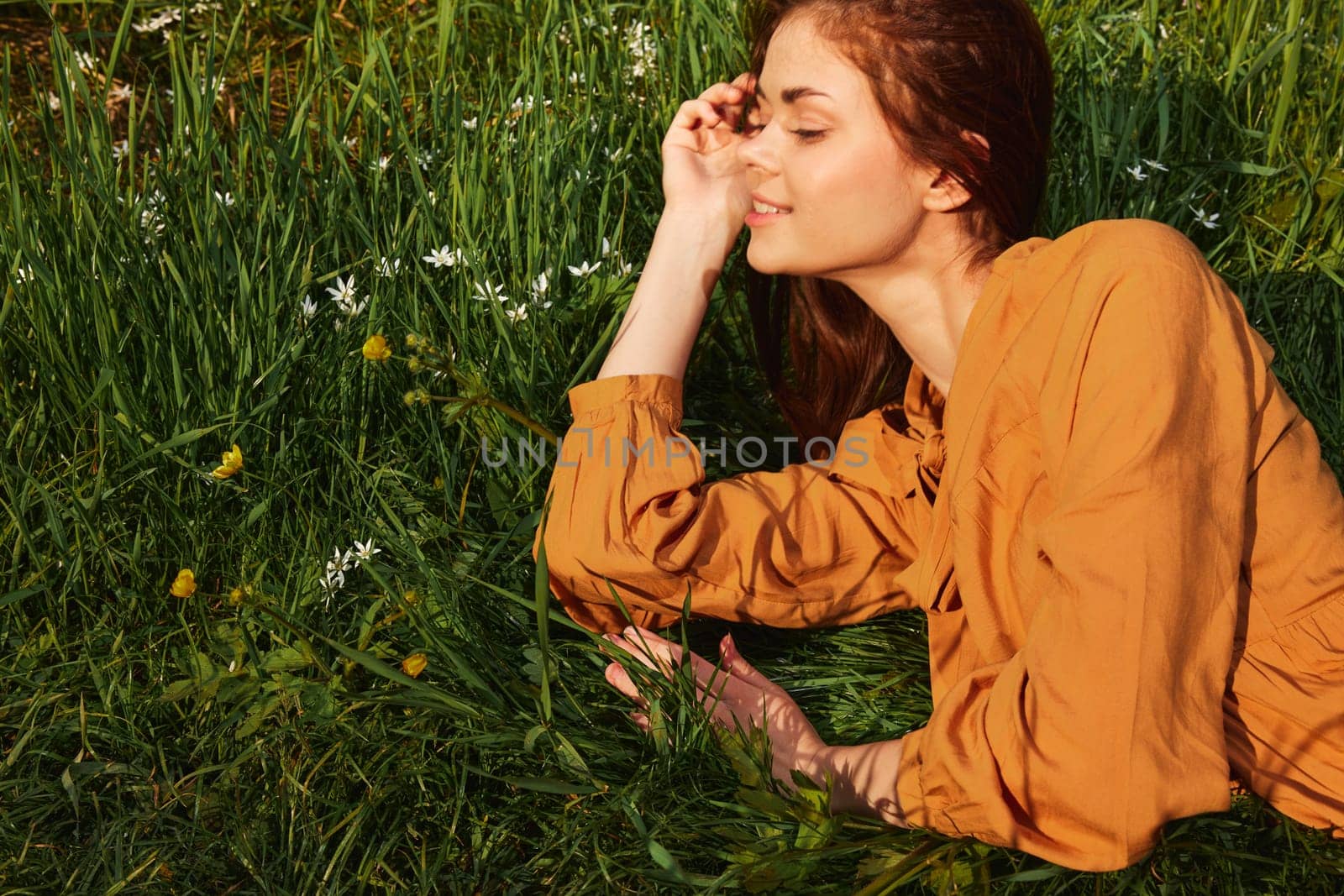 a calm woman with long red hair lies in a green field with yellow flowers, in an orange dress, smiling pleasantly, closing her eyes from the bright summer sun, resting her head on her hands. High quality photo