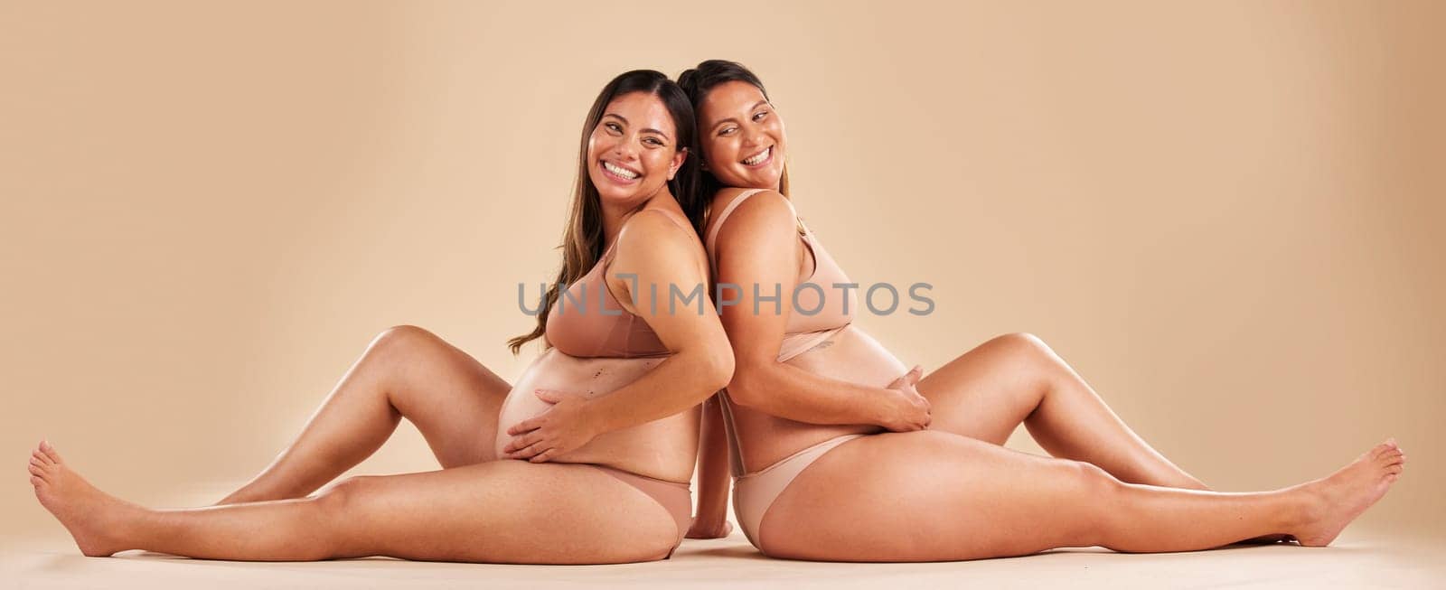 Pregnant woman friends, studio and underwear with smile, sitting and together for support by backdrop. Happy women, pregnancy and solidarity for wellness, care and holding healthy stomach by backdrop by YuriArcurs