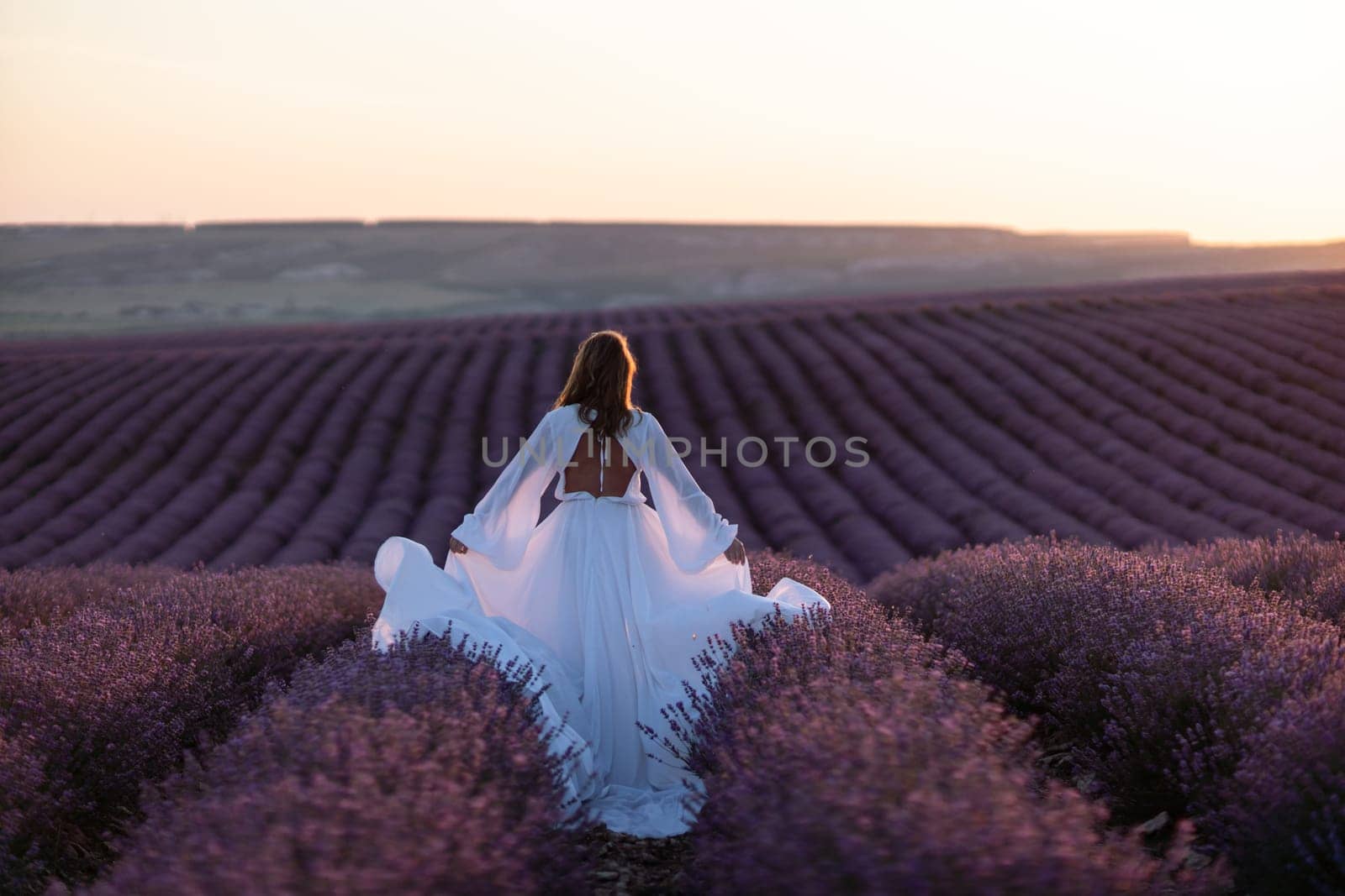 Happy woman in a white dress and straw hat strolling through a lavender field at sunrise, taking in the tranquil atmosphere