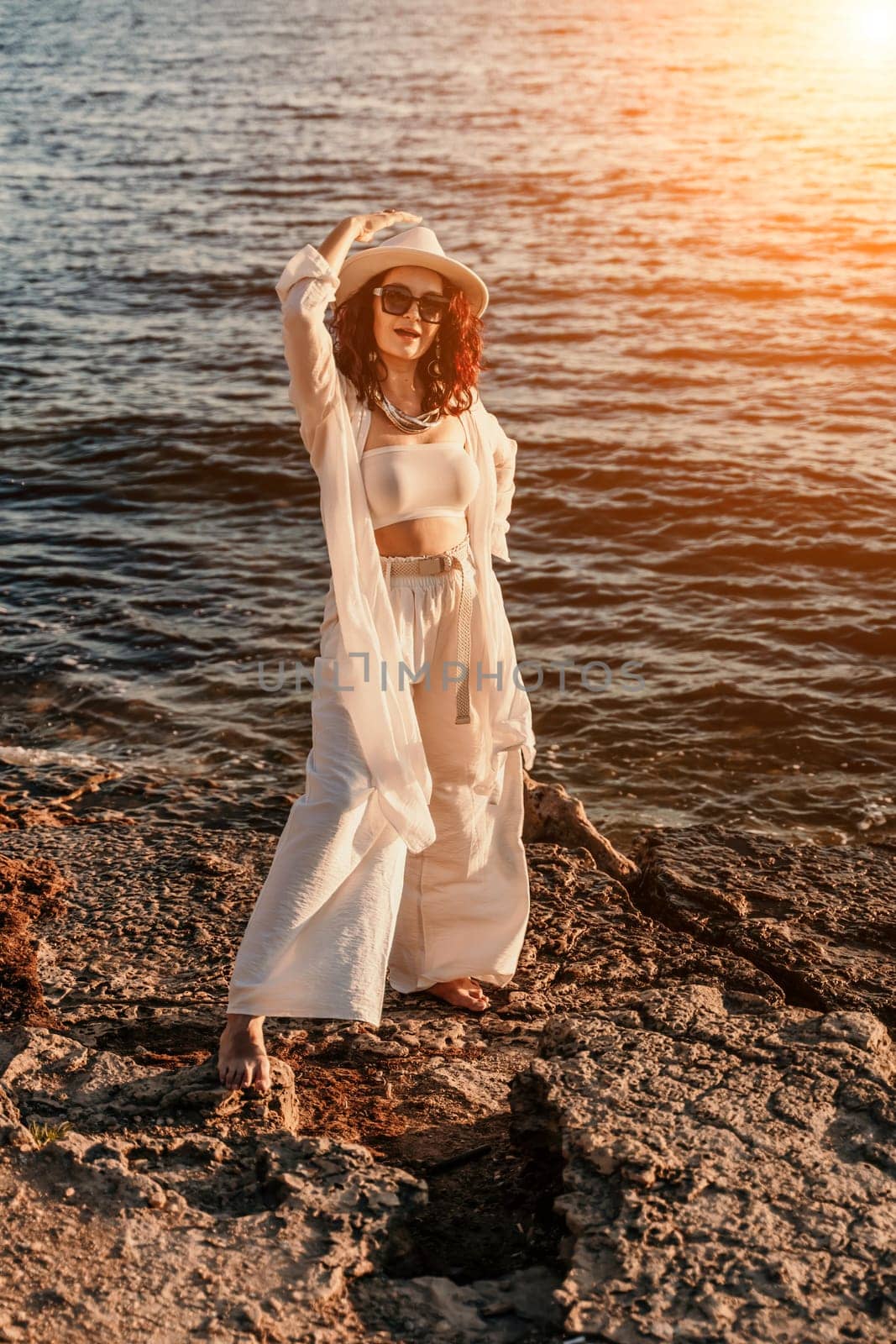 woman sea sunset. woman in a white pantsuit and hat is standing on the beach enjoying the sea. Happy summer holidays by Matiunina