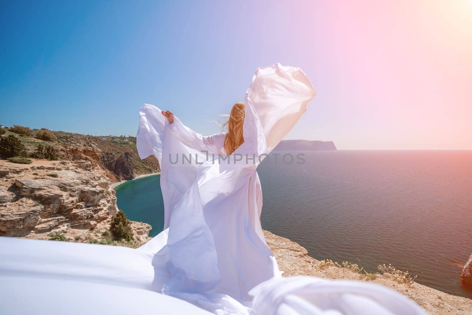 woman sea white dress. Blonde with long hair on a sunny seashore in a white flowing dress, rear view, silk fabric waving in the wind. Against the backdrop of the blue sky and mountains on the seashore