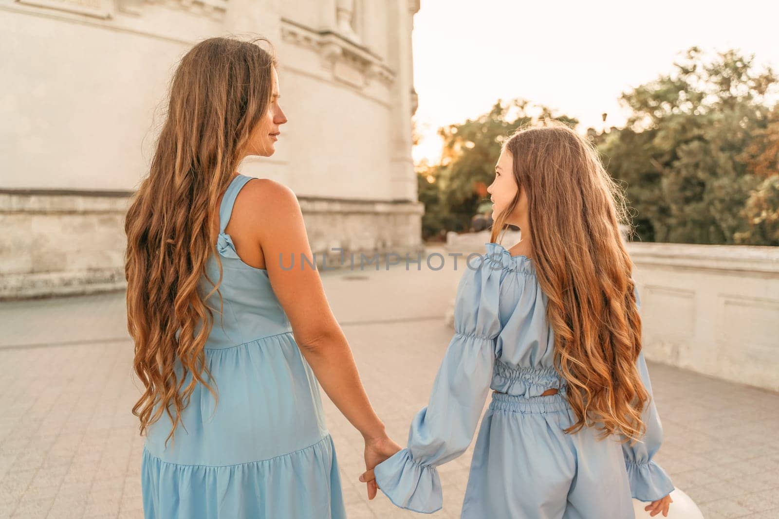 Daughter mother run holding hands. In blue dresses with flowing long hair, they hold balloons in their hands against the backdrop of a sunset and a white building. by Matiunina