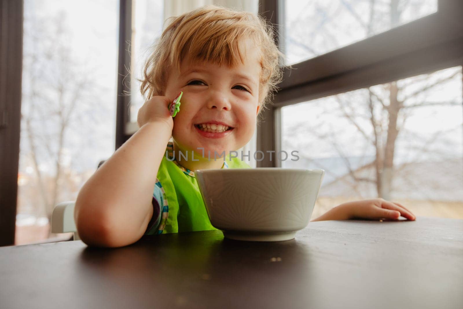 Smiling cute toddler sitting at table, eating and looking at camera indoors