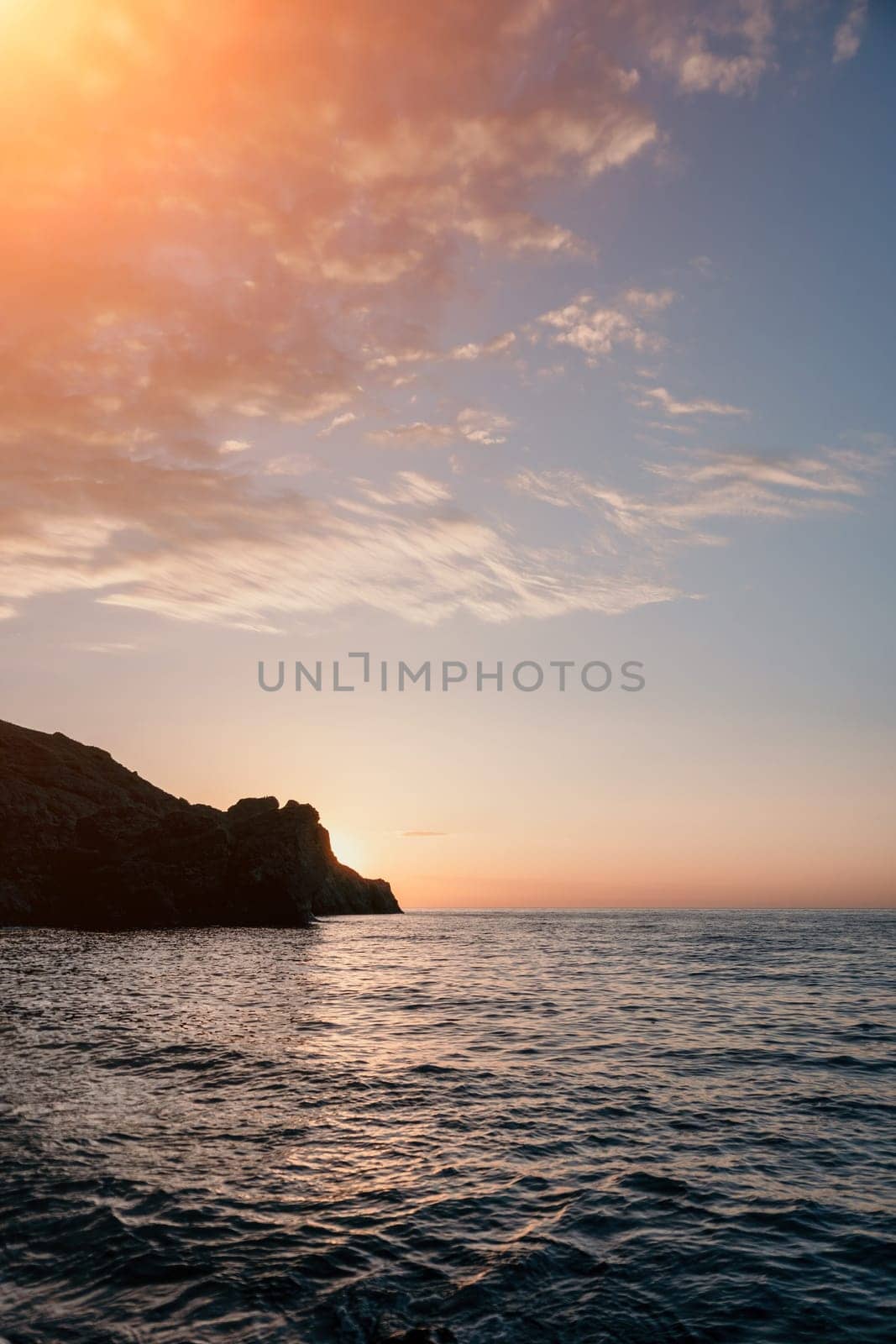 A red burning sunset with the silhouette of a cliff over the sea. Abstract nature summer or spring ocean sea background. Small waves on golden warm water surface with bokeh lights from sun.