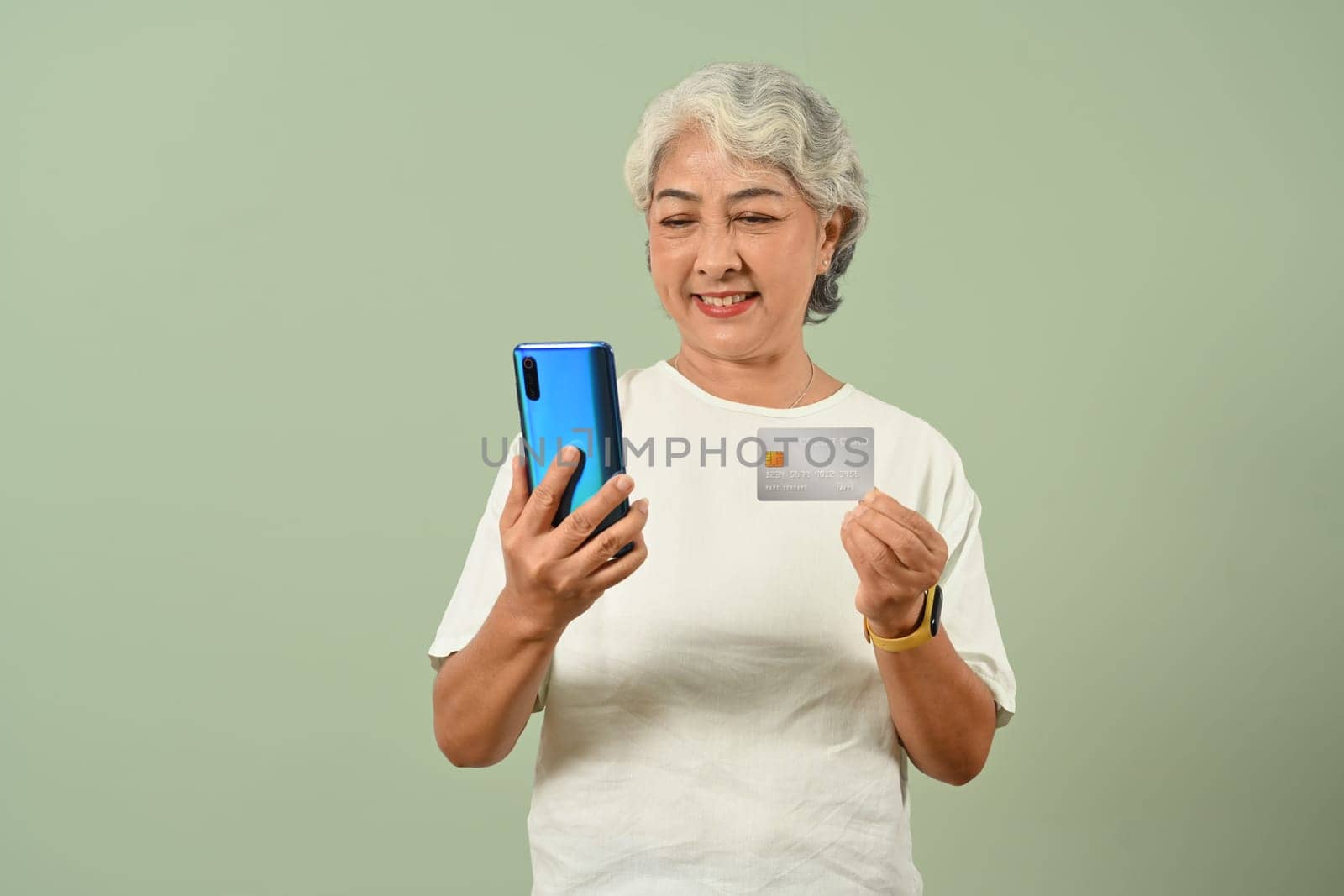 Satisfied mature woman 60s years with credit card using mobile phone to ordering in internet, standing over green pastel background by prathanchorruangsak