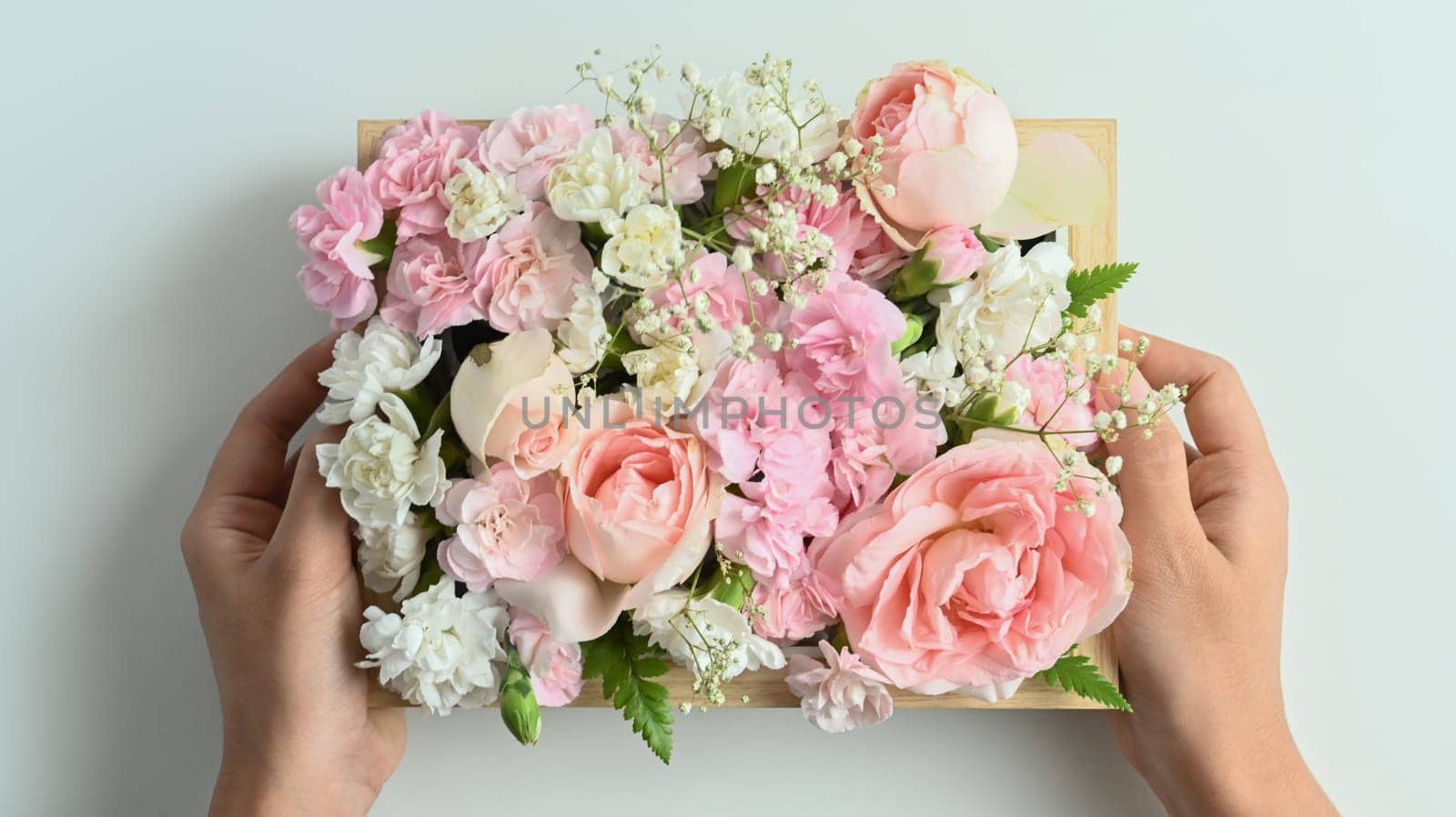Hands holding wooden crate full of pastel colors flowers with pink rose and carnation on white background.