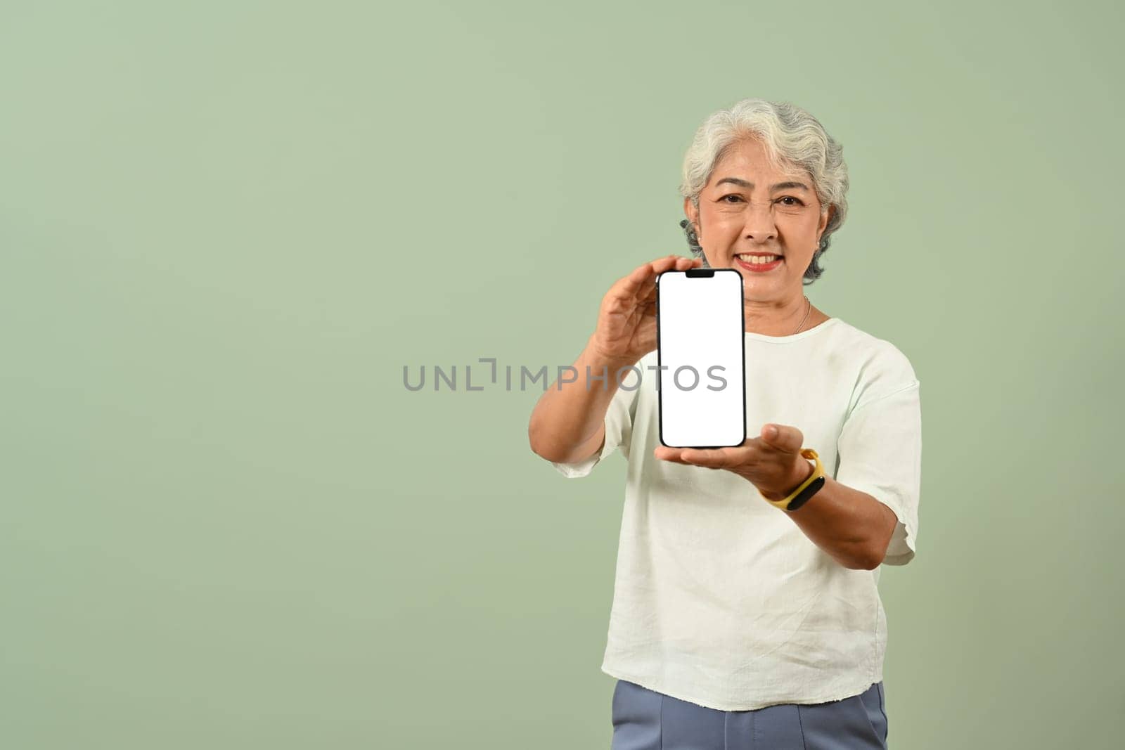 Positive senior gray haired woman showing smartphone with blank screen isolated on green background with space for text.