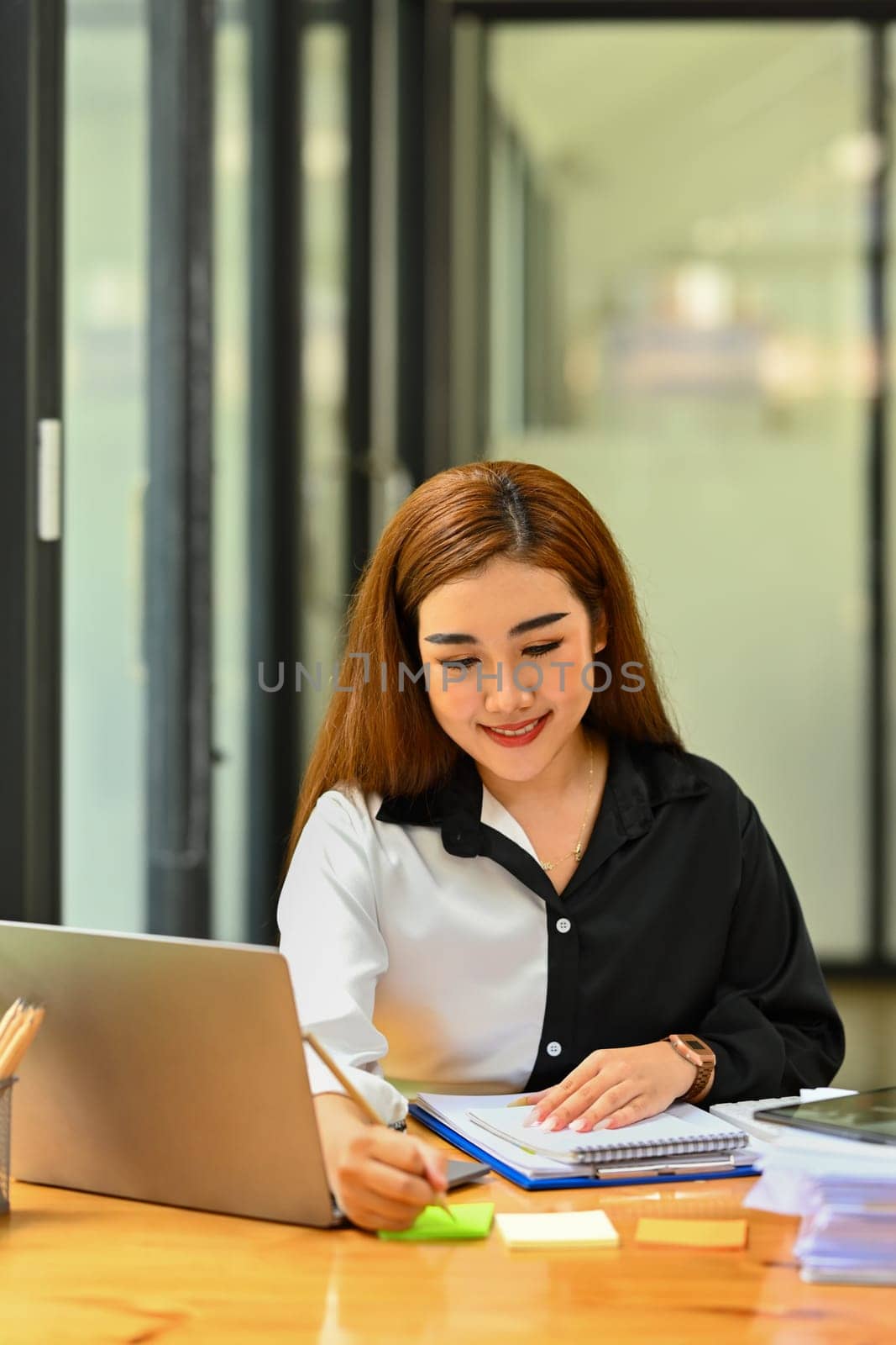Smiling female employee sitting in front of laptop and writing on sticky notes.