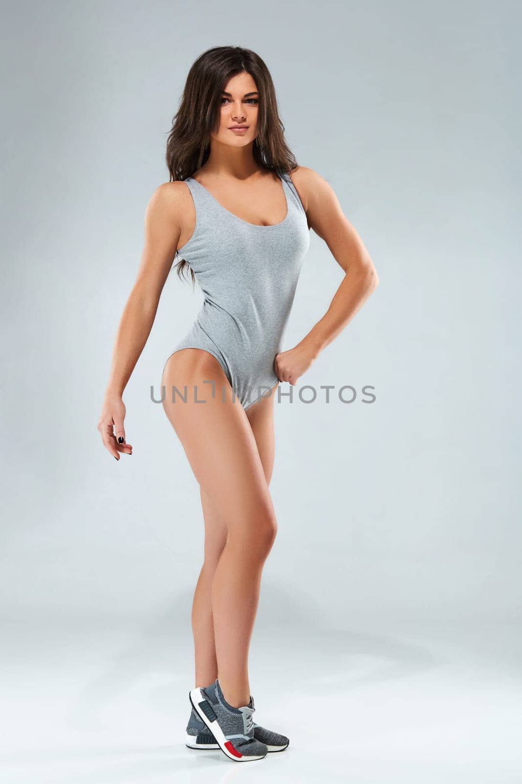 Fitness-instructor in sports clothing on gray background. Female model with fit muscular and slim body in sportswear. Young fit girl lifting dumbbells