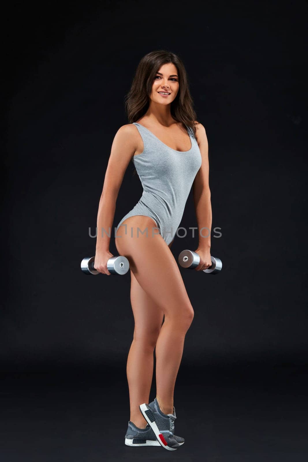 Fitness-instructor in sports clothing on black background. Female model with fit muscular and slim body in sportswear. Young fit girl lifting dumbbells