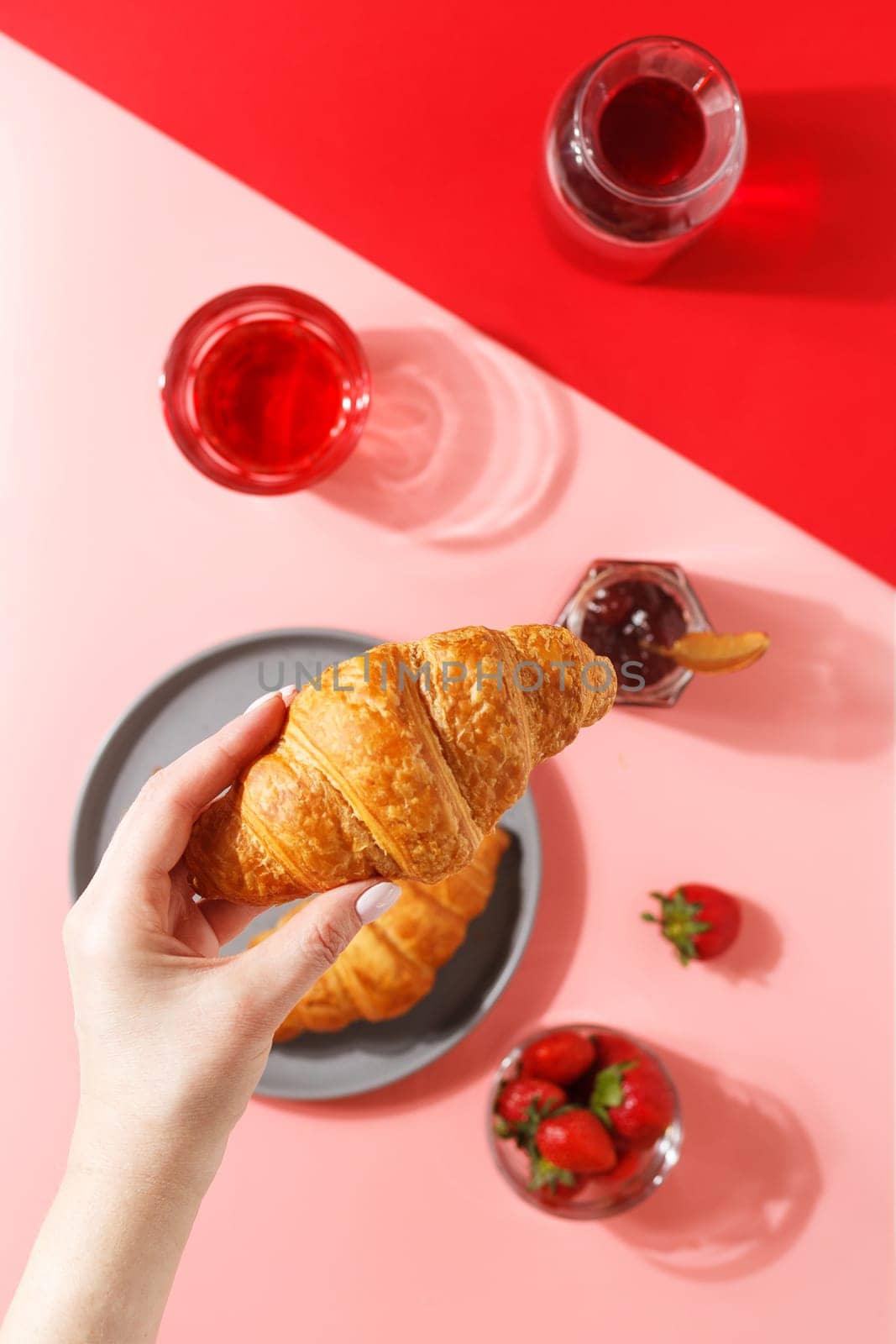 A woman's hand holds a kraussant over a plate with fresh puff croissants, berry juice, jam and fresh berries on a pink and red background.