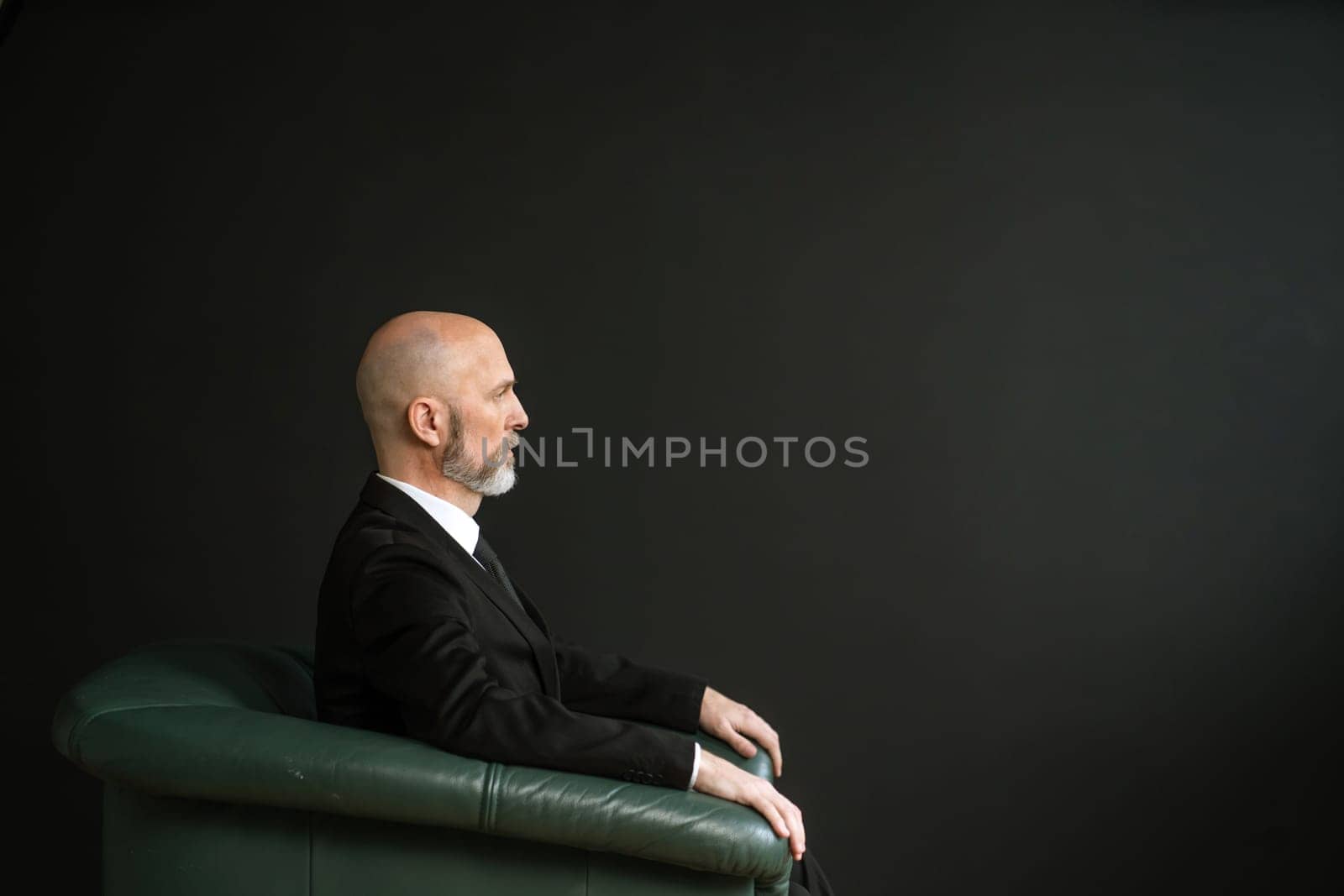 Mature man in a suit, sitting in a comfortable leather chair. The backdrop of a black wall adds a touch of elegance and sophistication to the image. High quality photo