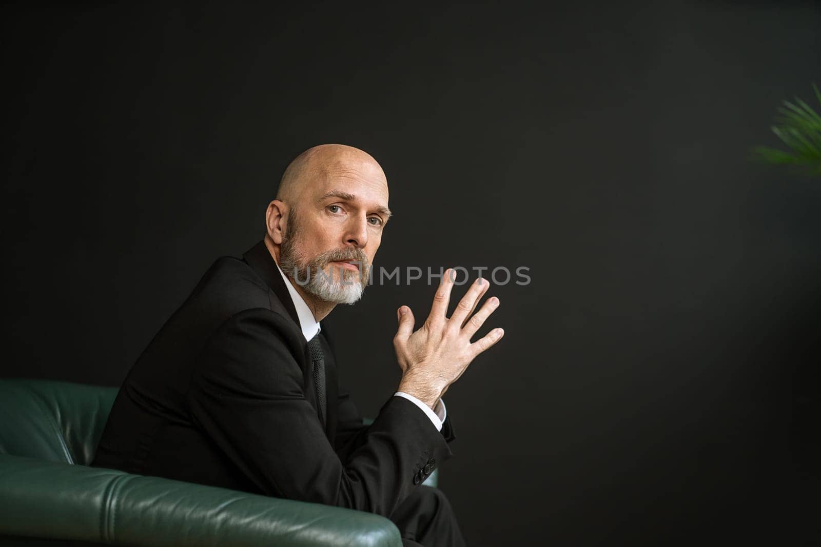 Solid and experienced elderly businessman with gray beard, he exudes confidence and professionalism. Seated in luxurious leather chair, he holds hands together in composed and thoughtful manner. by LipikStockMedia