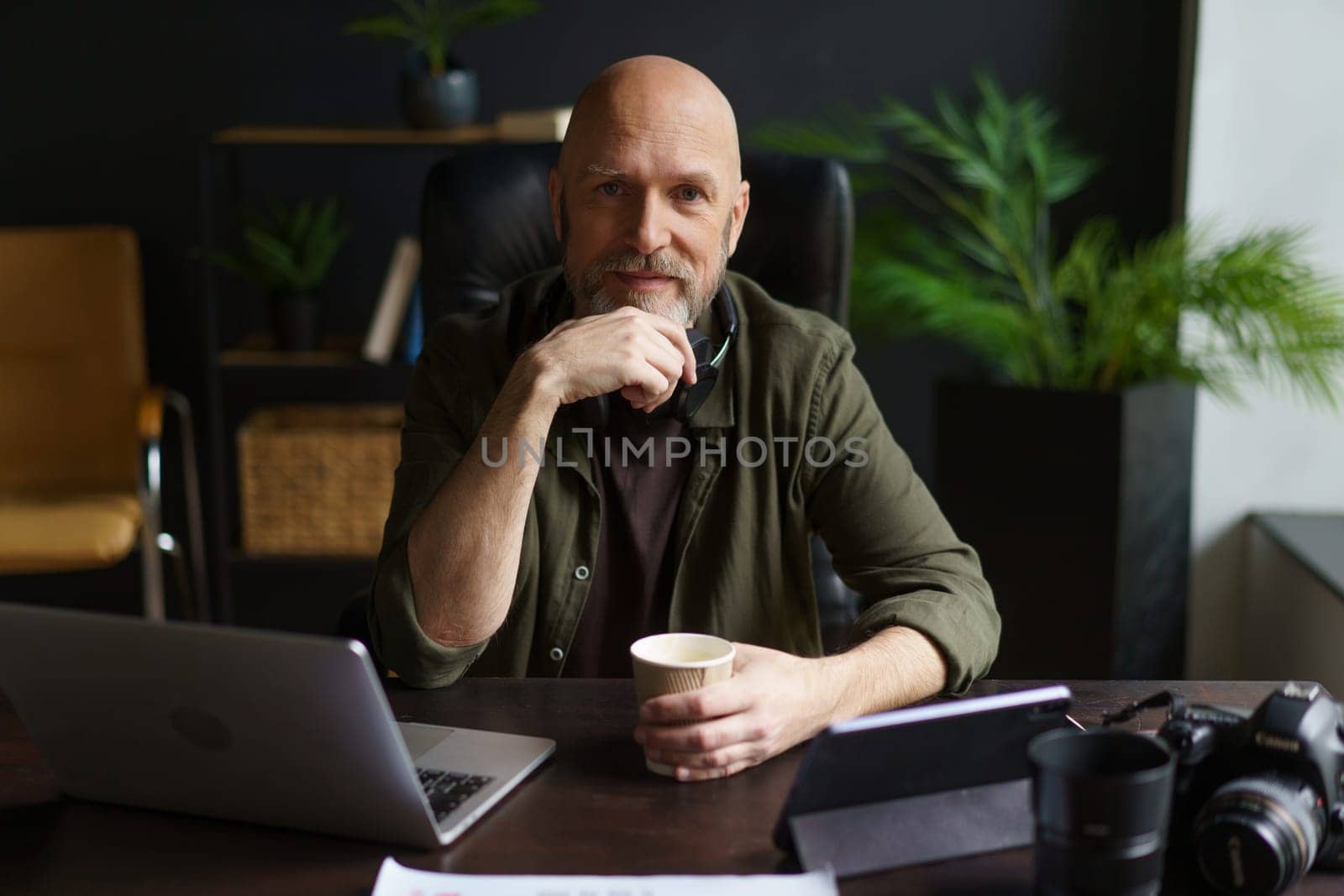 Old photographer, senior man, seated at desk in cozy home environment. table adorned with cup of coffee, photo camera, laptop, and various lenses, indicating deep involvement in world of photography. by LipikStockMedia
