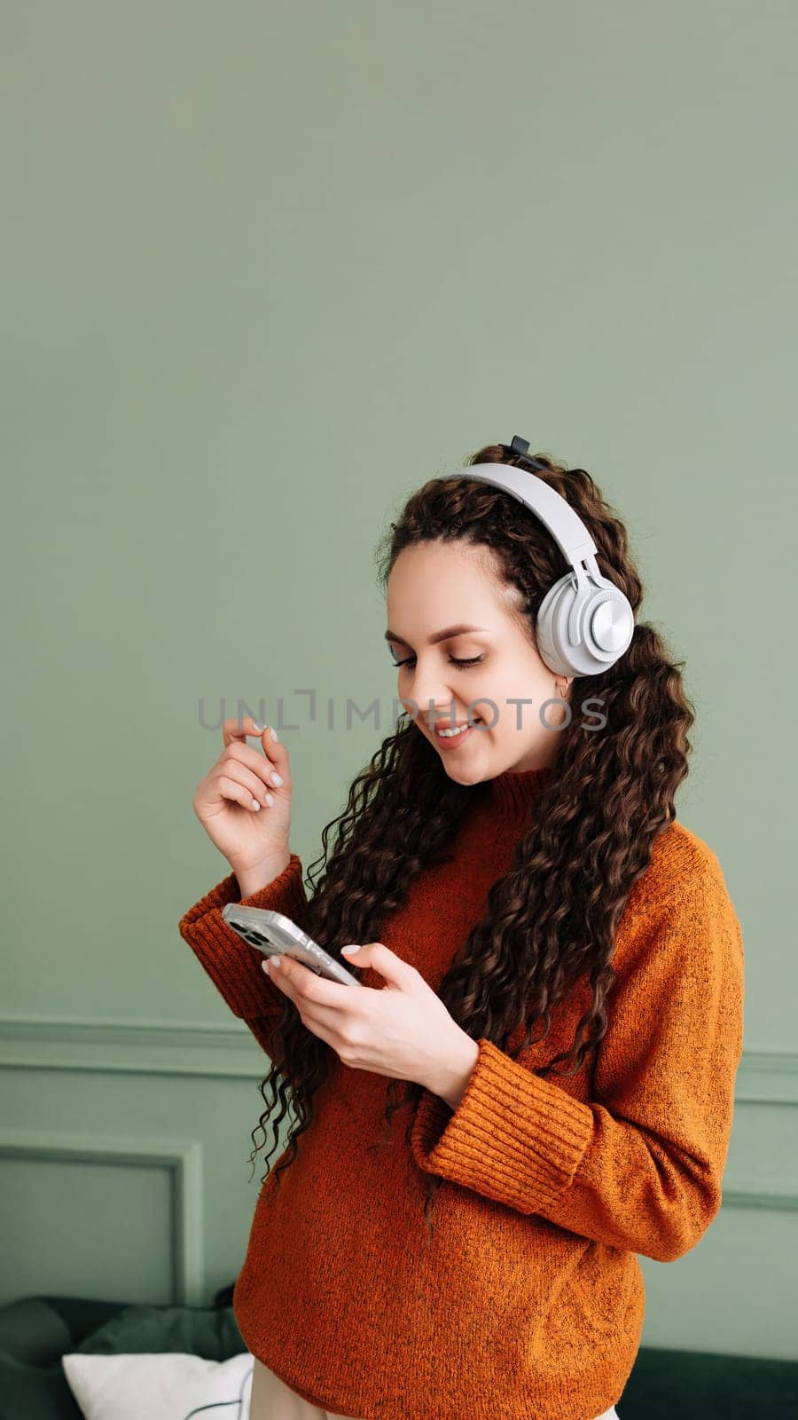 Happy pretty young woman wearing headphones dancing at home. Cheerful girl listening to music on mobile phone, singing songs feeling relaxed standing in modern living room interior