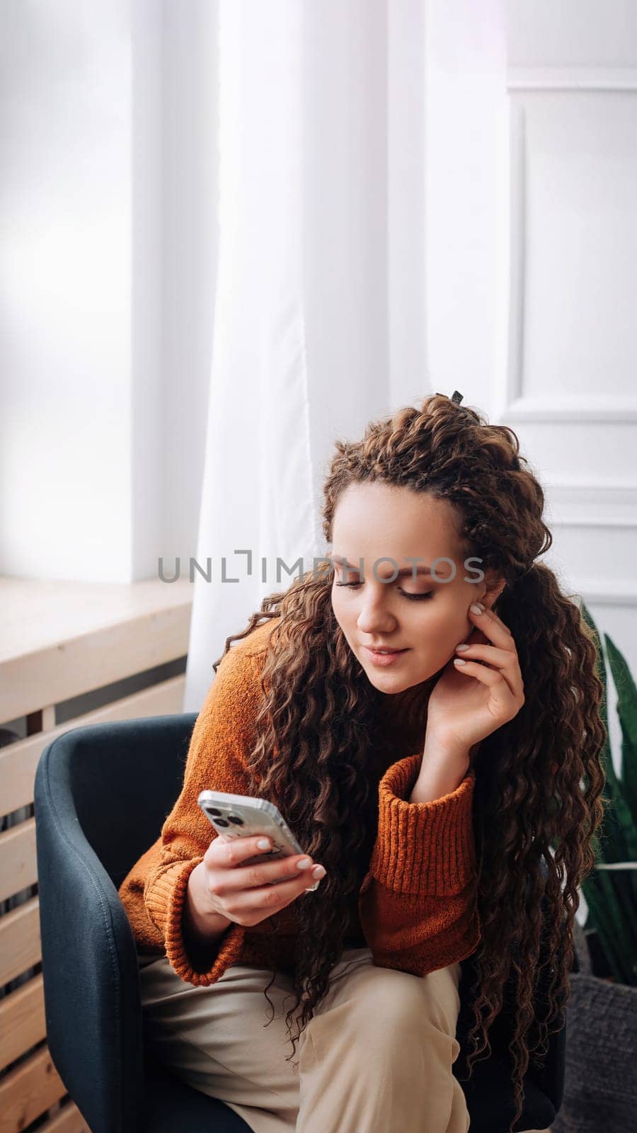 Smiling young woman sitting on chair holding mobile phone using cellphone device, looking at smartphone, checking modern apps, texting messages, browsing internet doing shopping relaxing at home