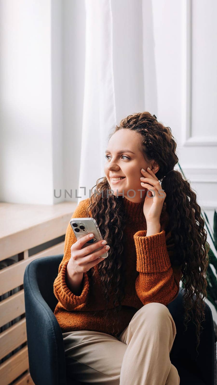 Young woman enjoying smartphone apps and texting while sitting on a chair. Mobile phone user: woman checking apps, messaging, and shopping from home. Relaxed woman browsing modern apps.