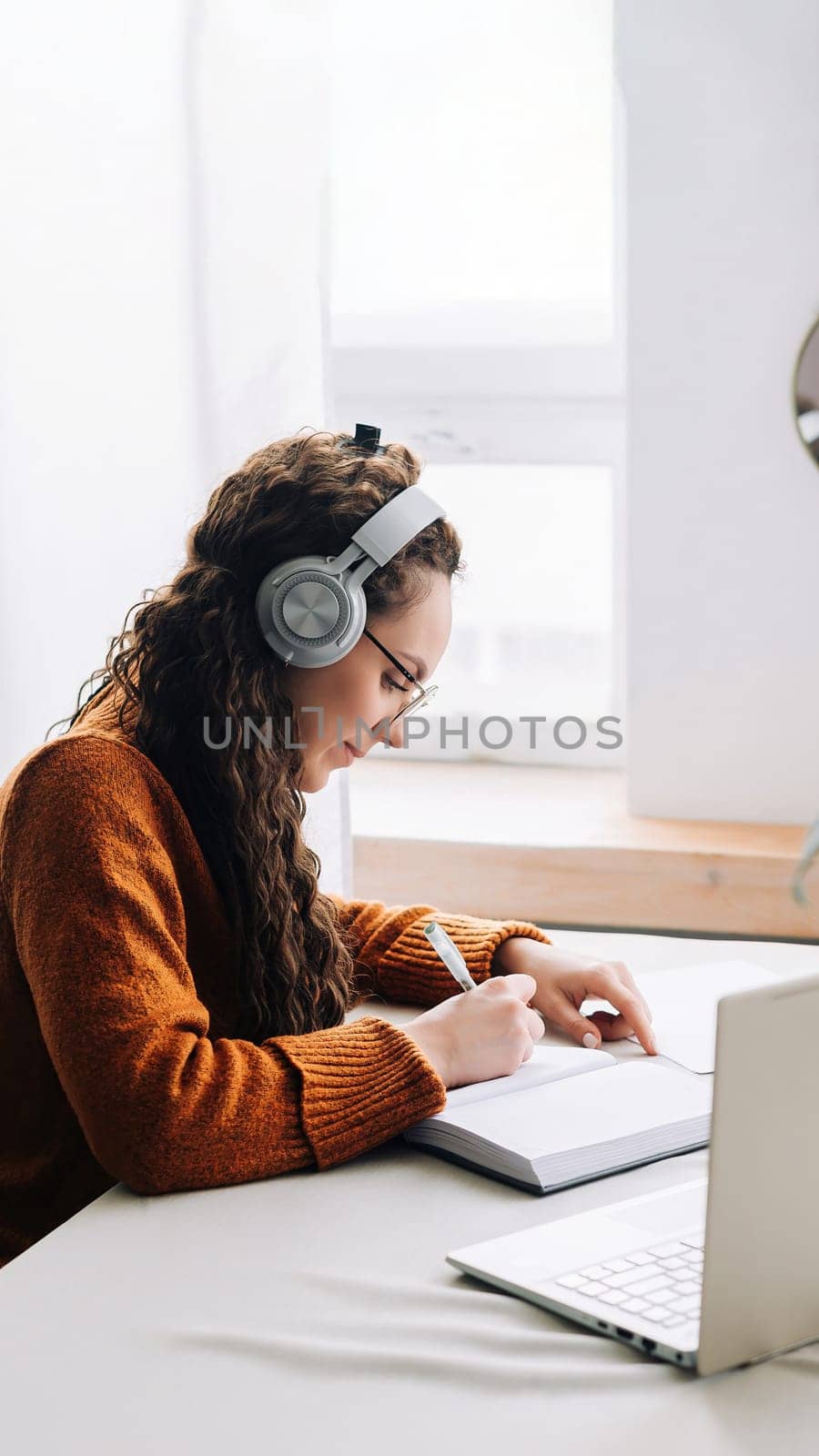 Busy young woman university student wearing headphones using laptop e-learning, writing notes, studying online education seminar class by webinar, learning online seminar sitting at the home table