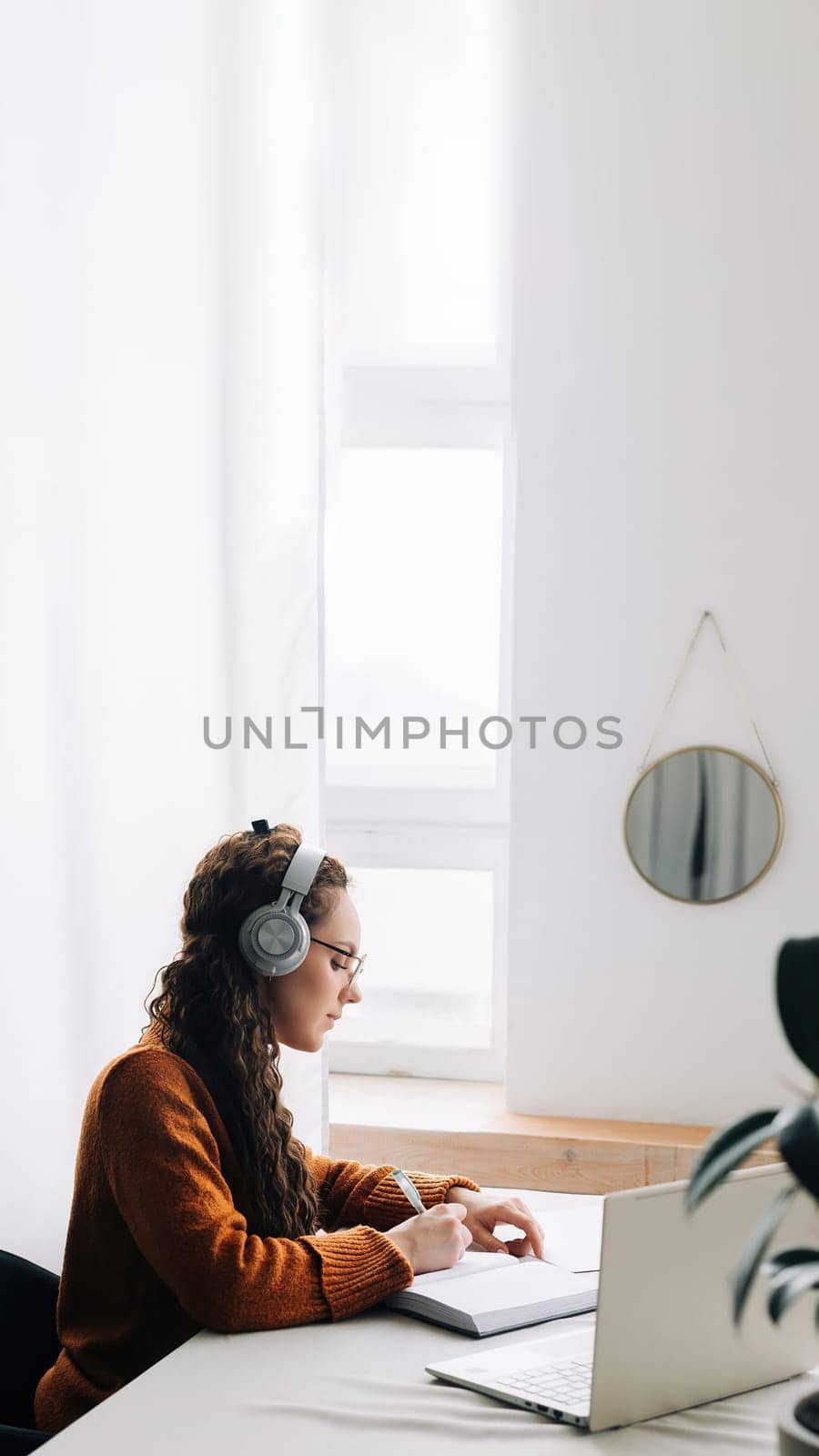 Young student attending virtual seminar while studying at home. Woman wearing headphones and typing on laptop, taking notes during e-learning session or online class