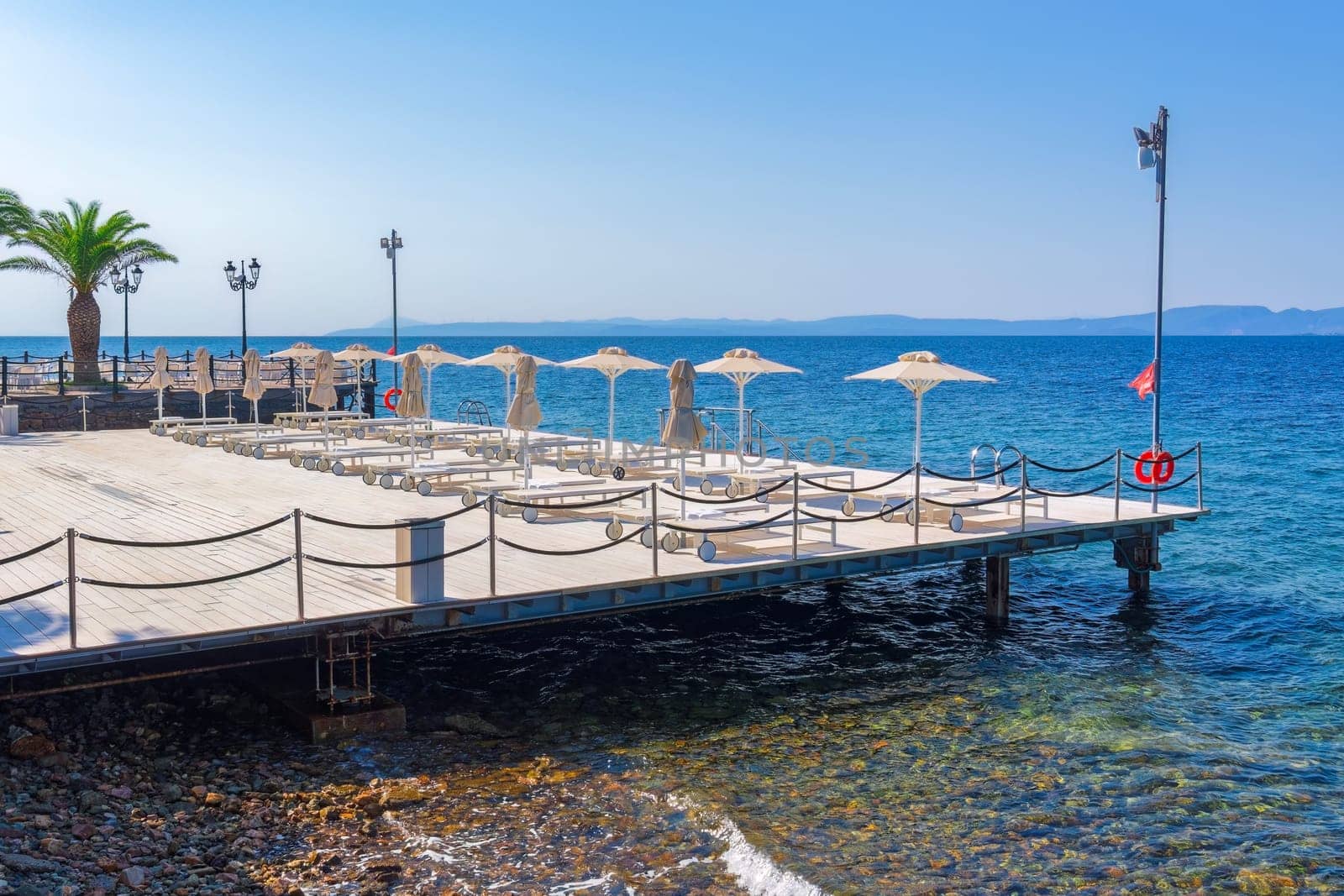 Empty wooden sunbathing deck with chairs and umbrellas by a calm blue sea under the bright sun in summer.