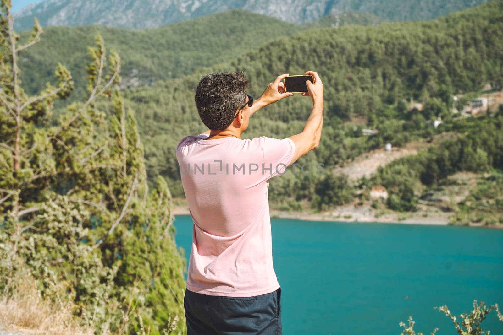 Man shooting a video on mobile phone of mountains lake background. Traveler male taking photo on cellphone on the blue lake outdoors travel adventure vacation.