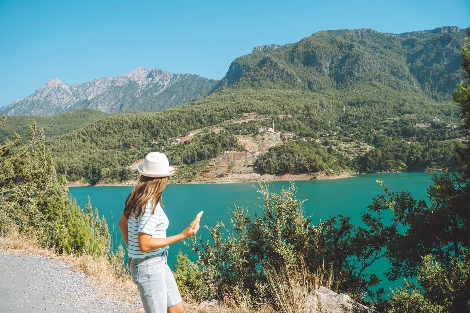 Smiling woman in hat and sunglasses with wild hair standing near mountains lake on background. Positive young woman traveling on blue lake outdoors travel adventure vacation.