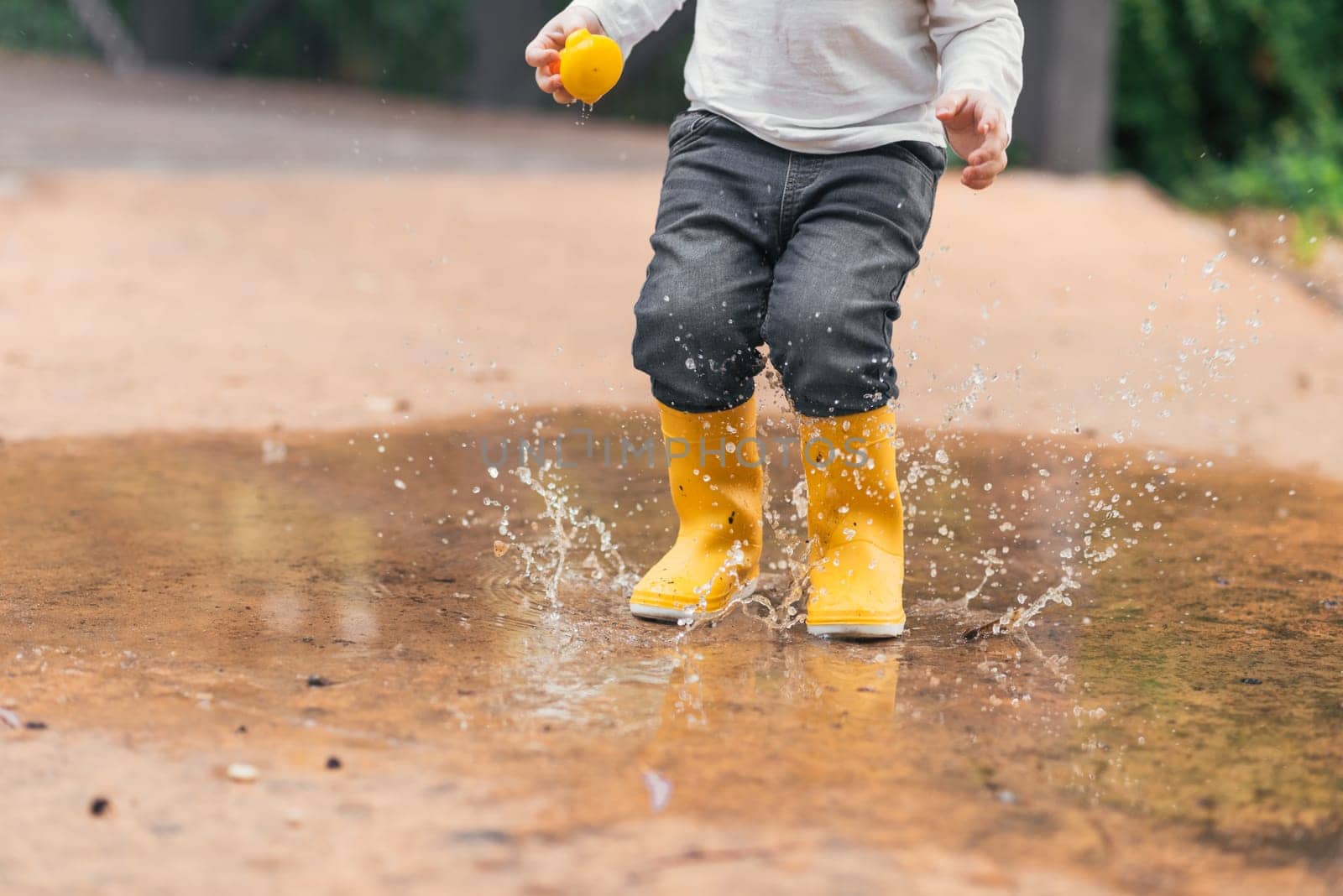 Child's feet in yellow rubber boots jumping over a puddle in the rain.