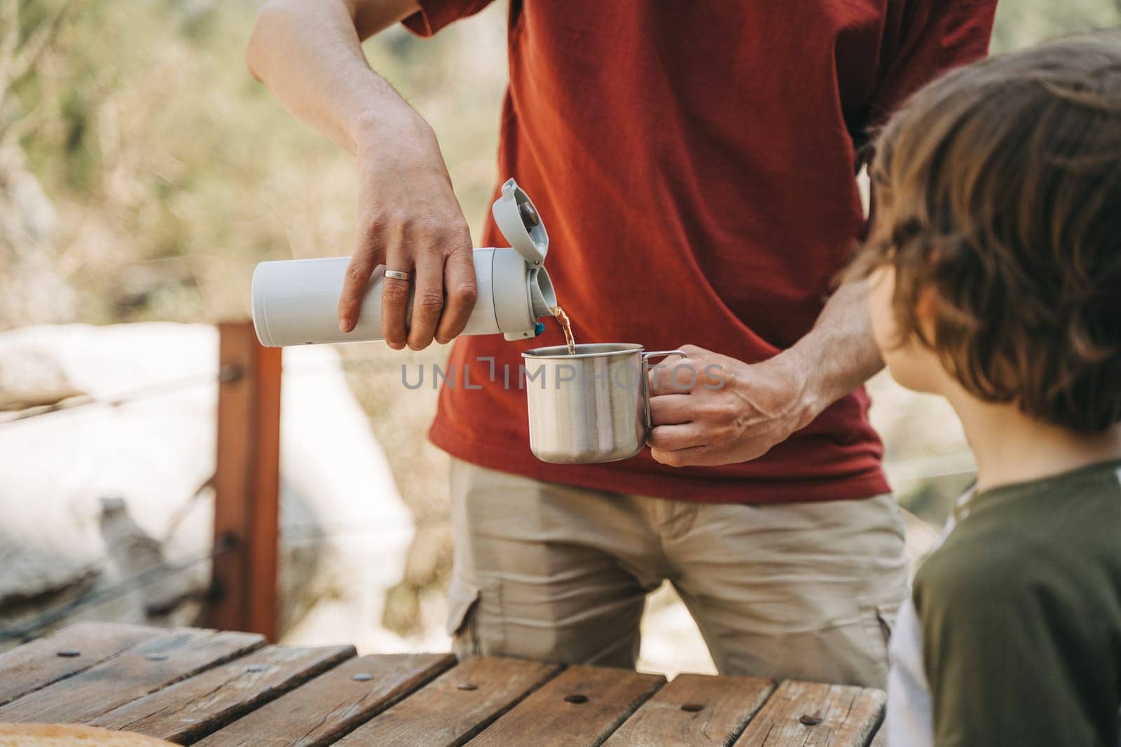 Father dad pours hot coffee tea from thermos into the mug on a family picnic in the mountains. Child school boy kid is watching his dad filling the cup with hot beverage by Ostanina