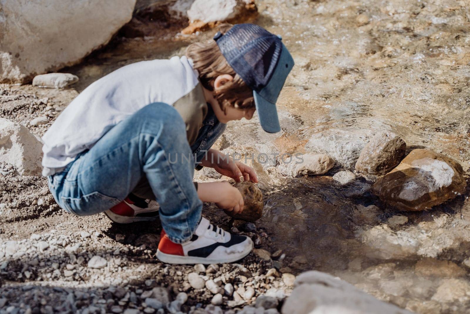 Caucasian school boy throwing rocks into the canyon river. Kid child playing with stones while sitting on a boulder near mountain river bank.