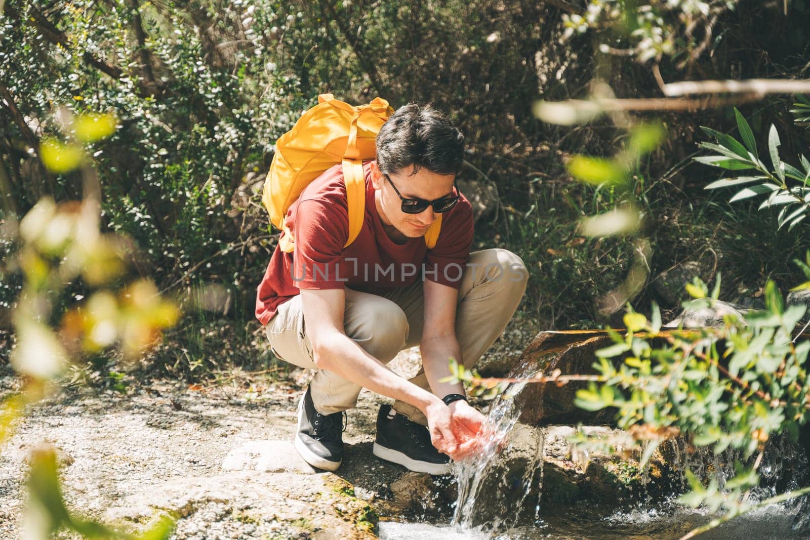 Adult man drinking water from the mountain creek. Tourist wearing casual clothes and sunglasses making a sip of mountain river water from the palms of his hands when hiking.