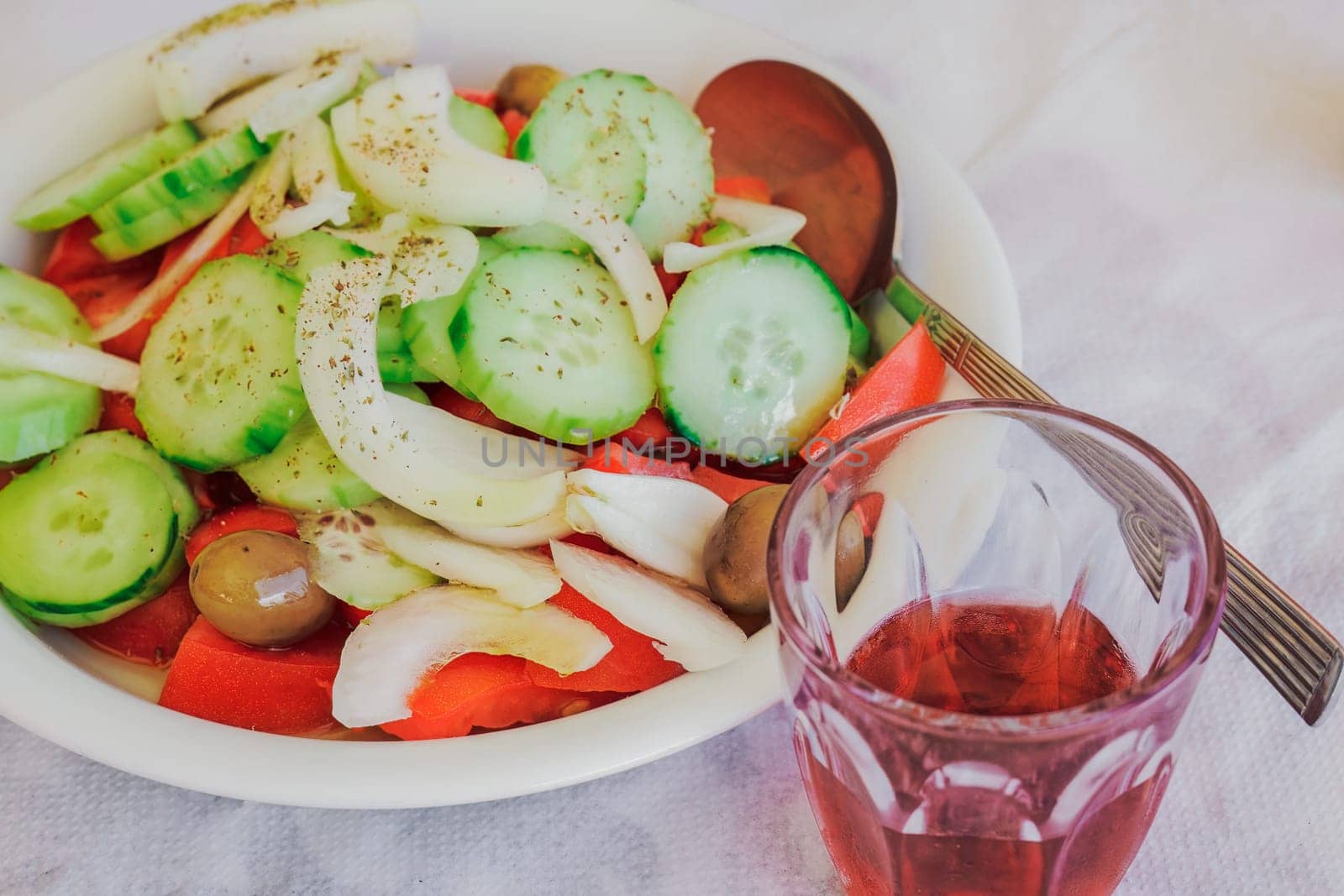 A small glass of red wine next to a dish of served salad with sliced cucumber, tomatoes, onions and olives.