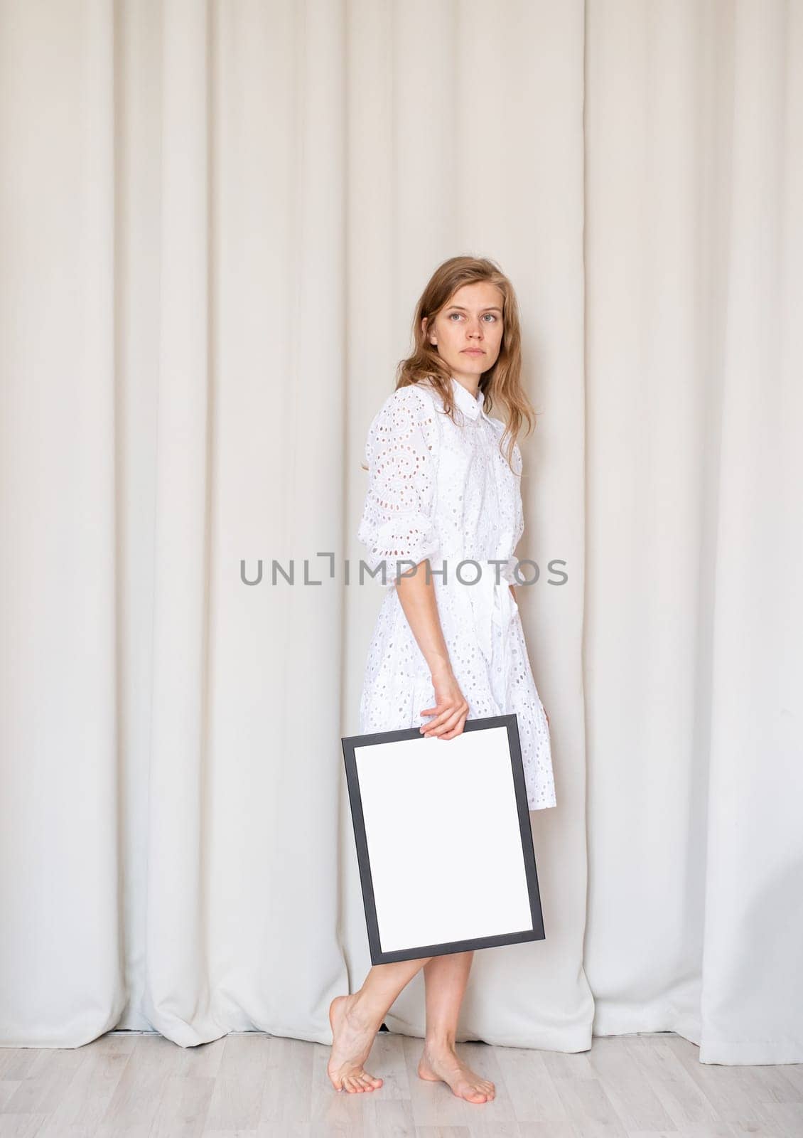 woman in dress holding blank frame, beige curtain background by Desperada